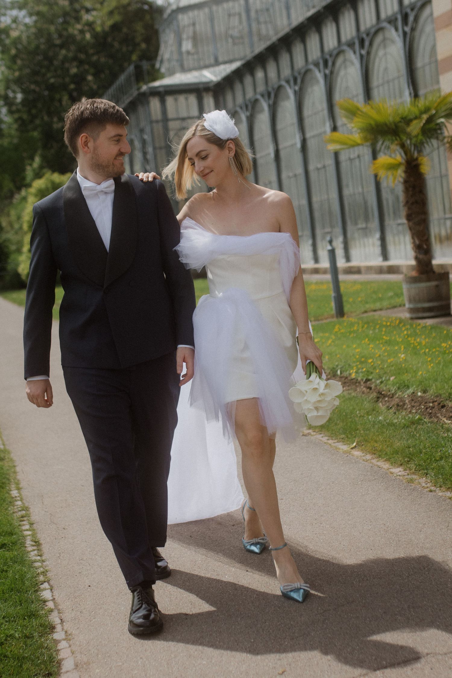 Newlyweds walking together on garden path, bride wearing short wedding dress with flowing train.