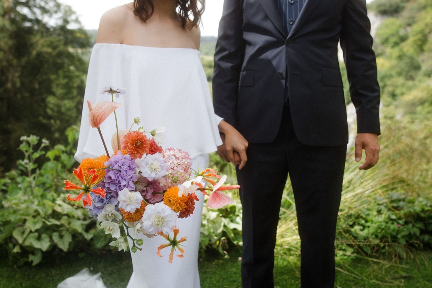 A close-up of a wedding bouquet with orange and purple flowers against white dress and dark suit.