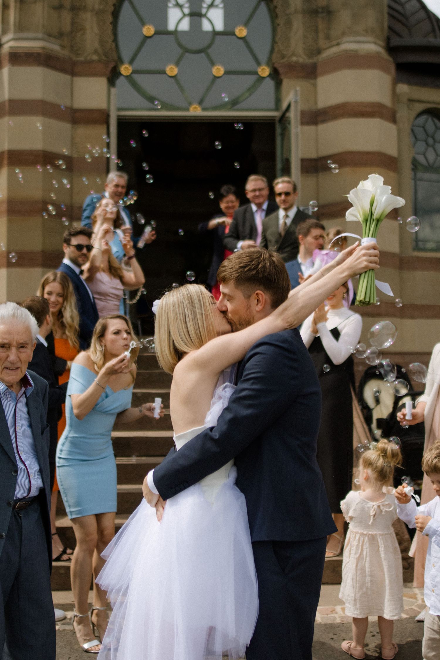 Wedding couple sharing romantic moment outside church while guests blow bubbles in celebration.