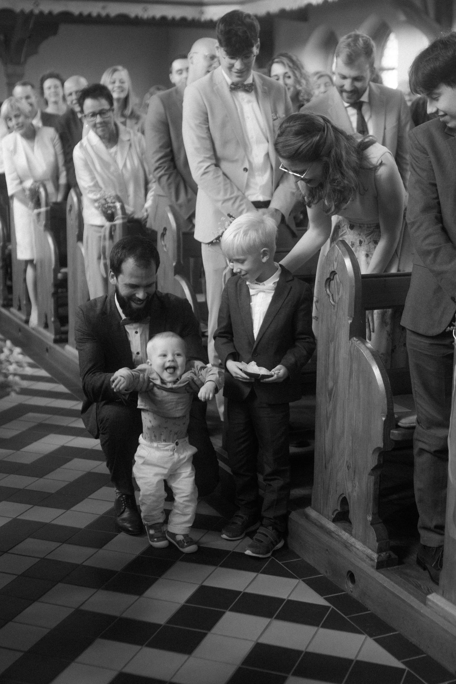 Wedding guests gather around small children in church with black and white checkerboard floor.