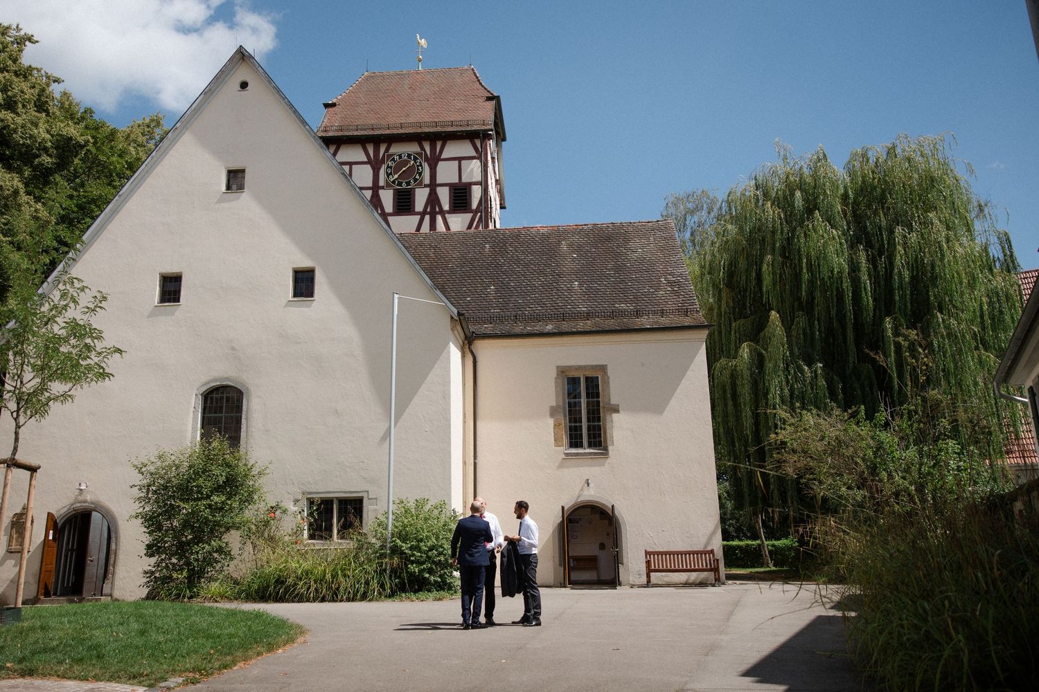 Historic white church building with timber-framed tower and weeping willow tree on a sunny day.