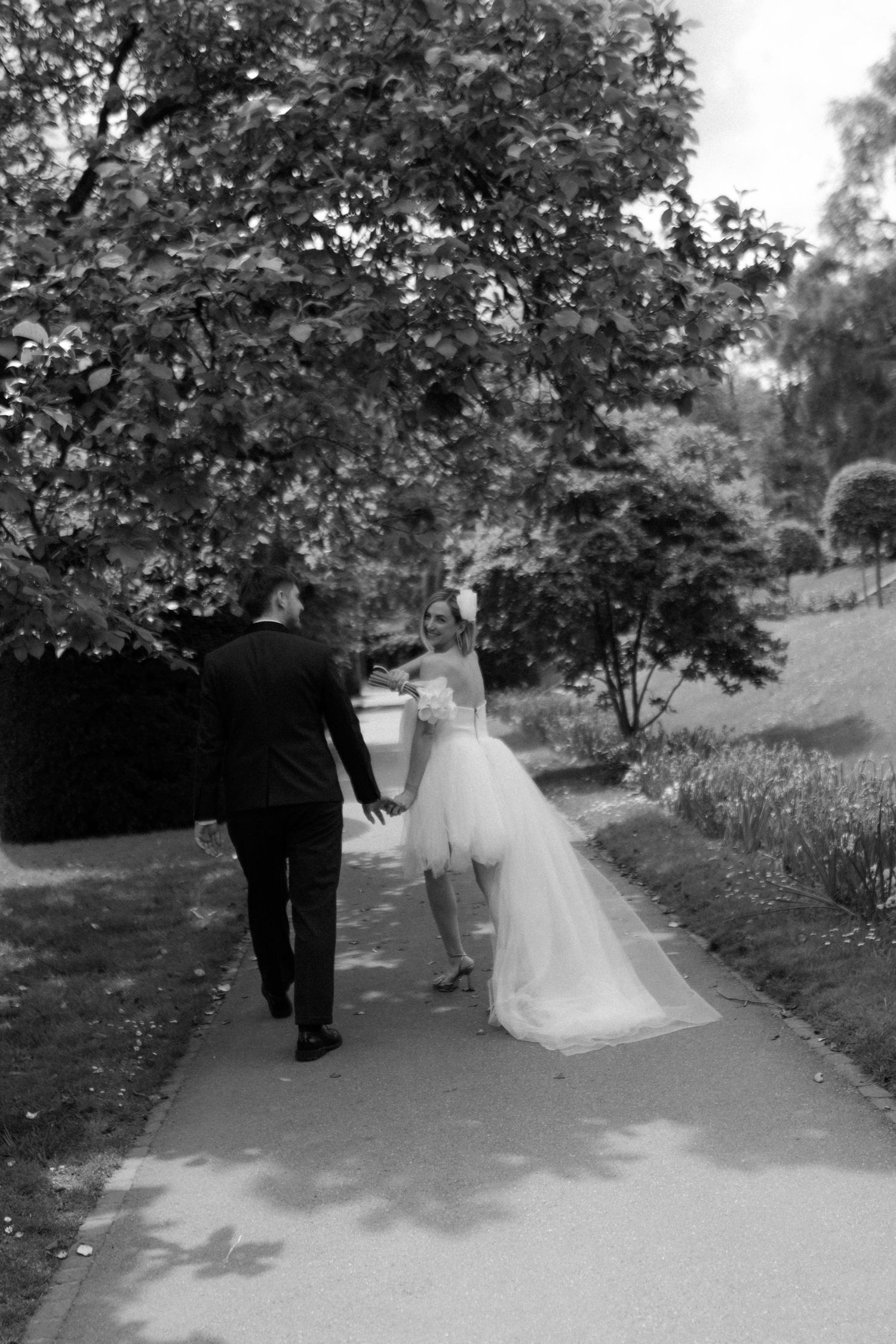 Black and white wedding photograph capturing a couple strolling down a tree-lined garden path.