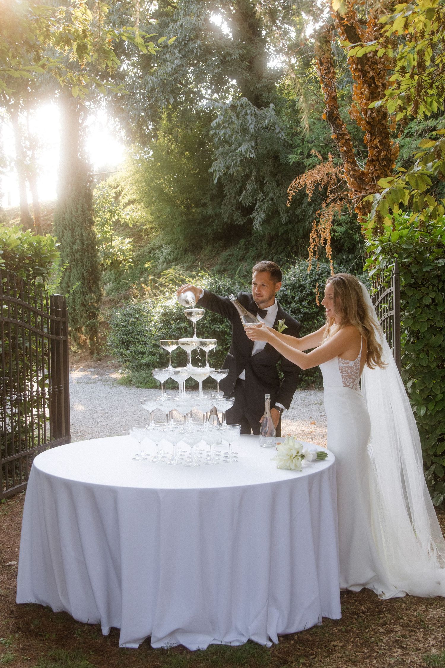 Bride and groom carefully pouring champagne into a pyramid of glasses in a garden setting.