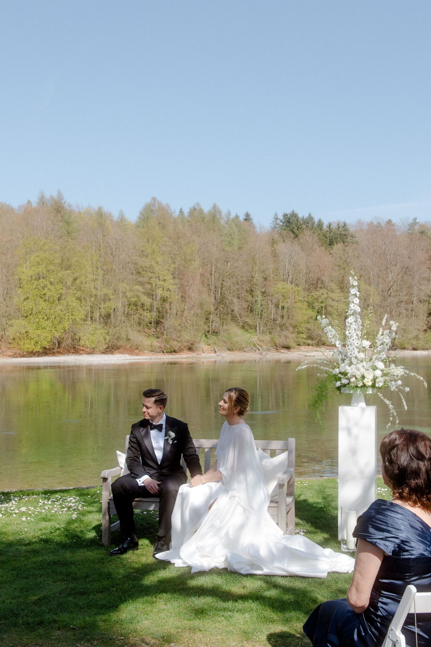 Bride and groom sit together at lakeside outdoor wedding ceremony with white floral arrangements.