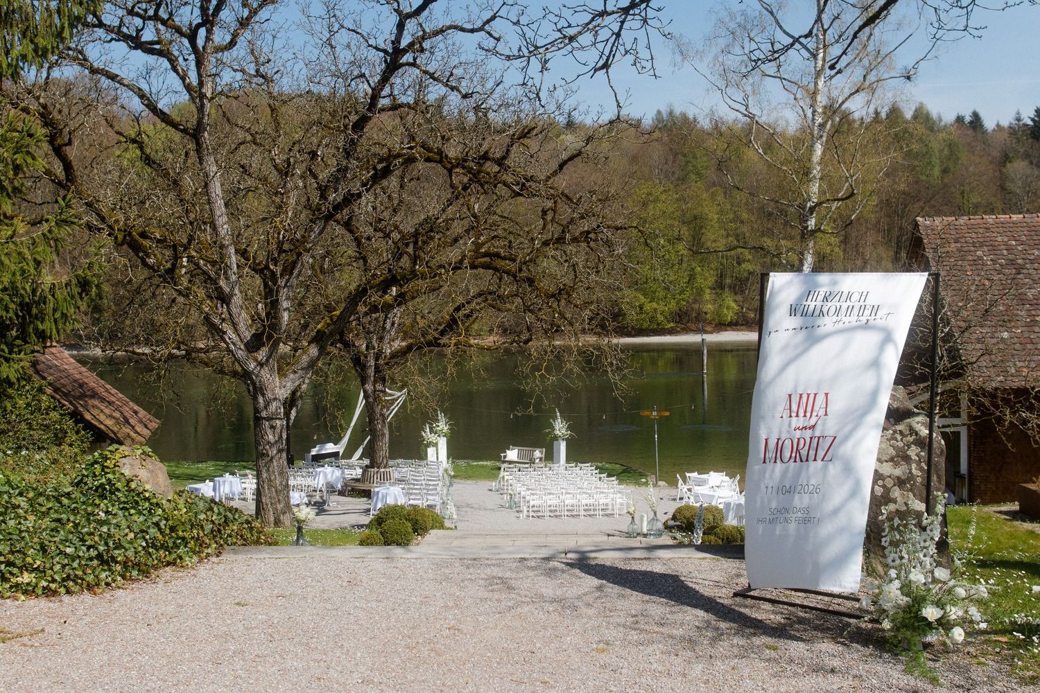 Scenic lakeside venue with white chairs set for an outdoor event, surrounded by trees and a large decorative sign.
