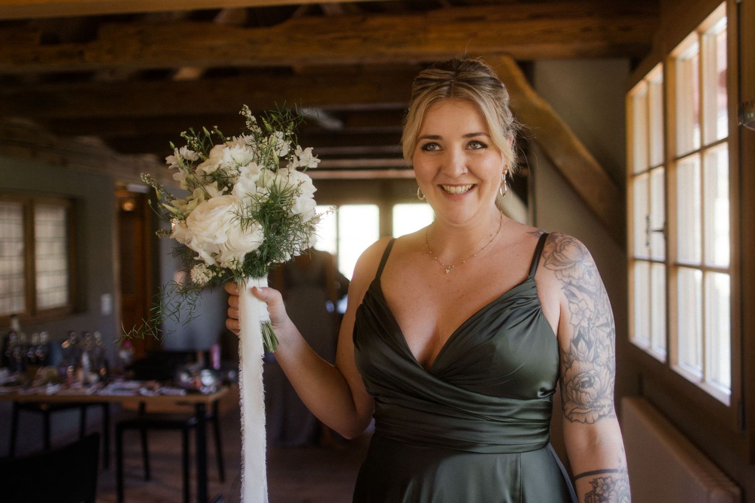 Smiling bridesmaid in olive green gown holding white floral bouquet with visible arm tattoos in rustic wedding venue.