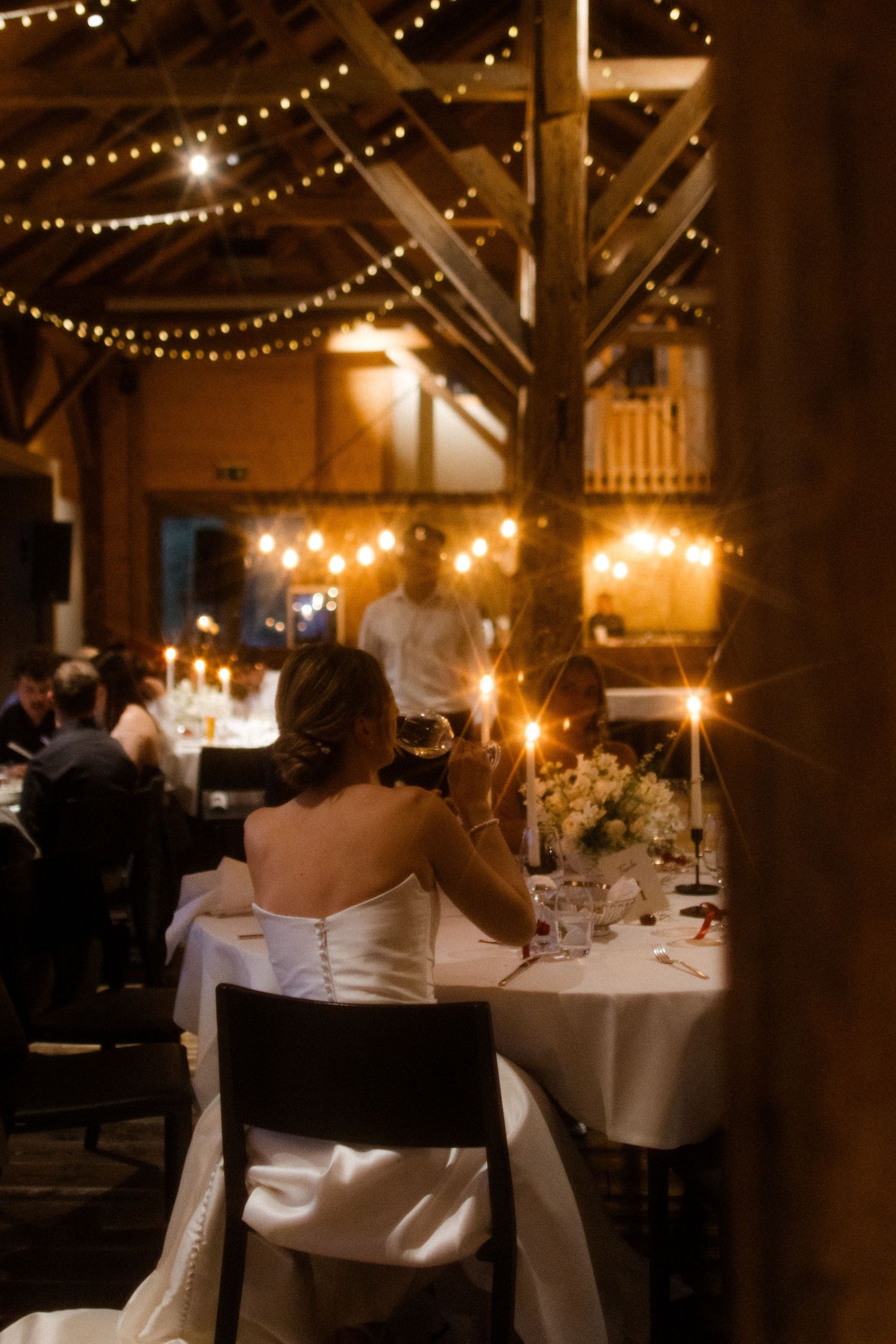 Bride in white strapless gown seated at candlelit wedding reception table in rustic barn with string lights.