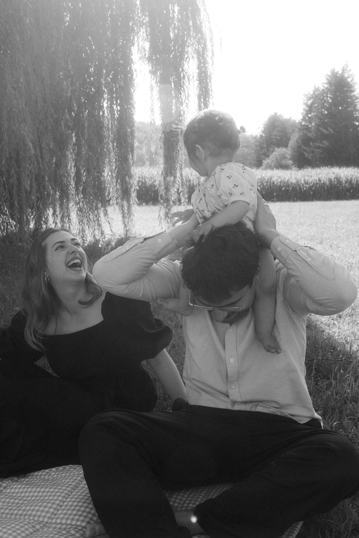 Black and white photo of a laughing couple sitting outdoors, playfully lifting their baby up in the air.