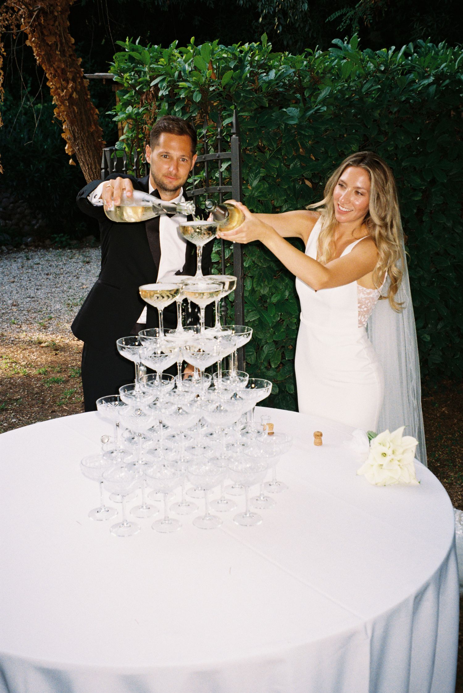 A couple pours champagne into a tower of crystal glasses at an elegant outdoor wedding reception.