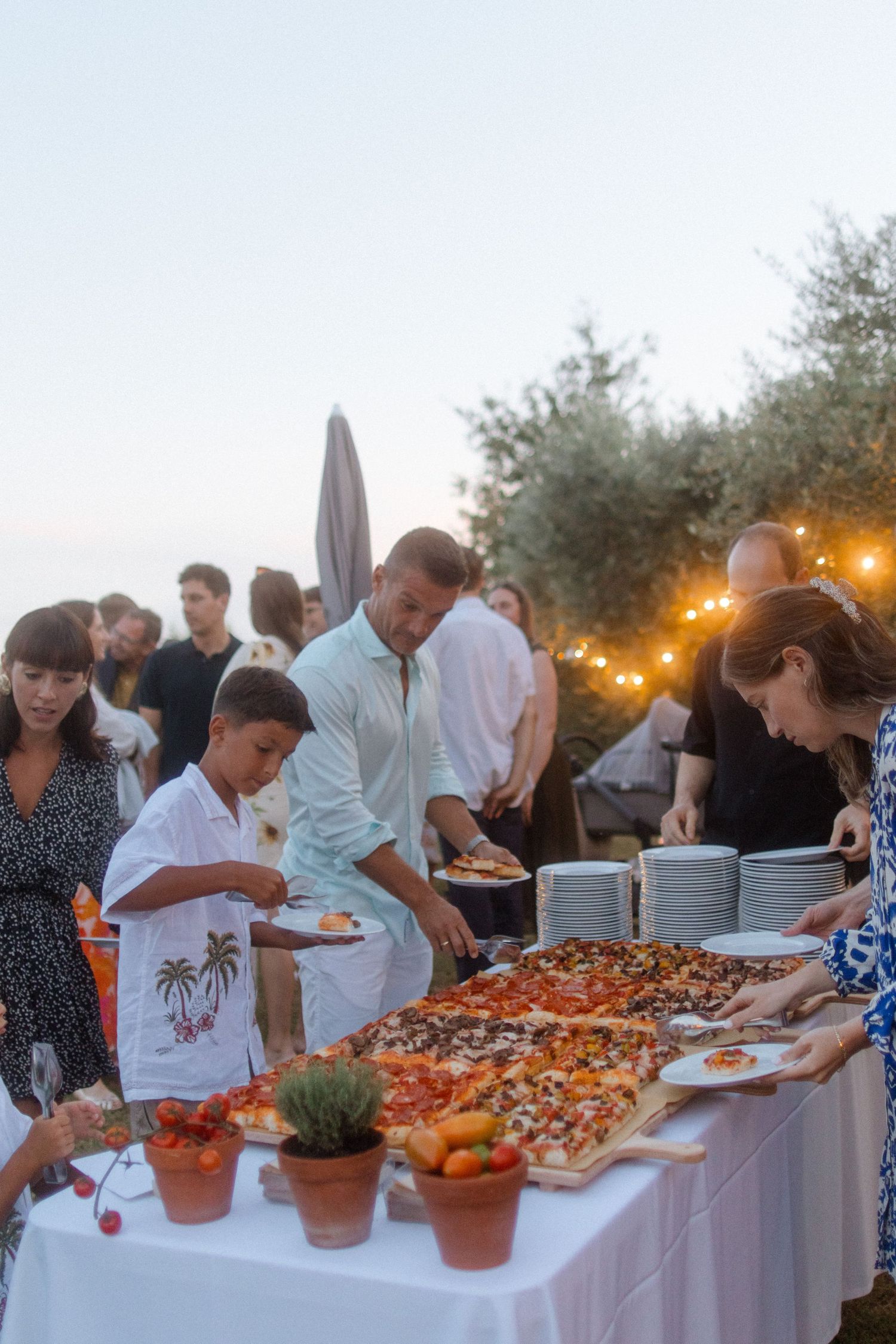 People gathering around a buffet table with food platters under string lights at dusk.
