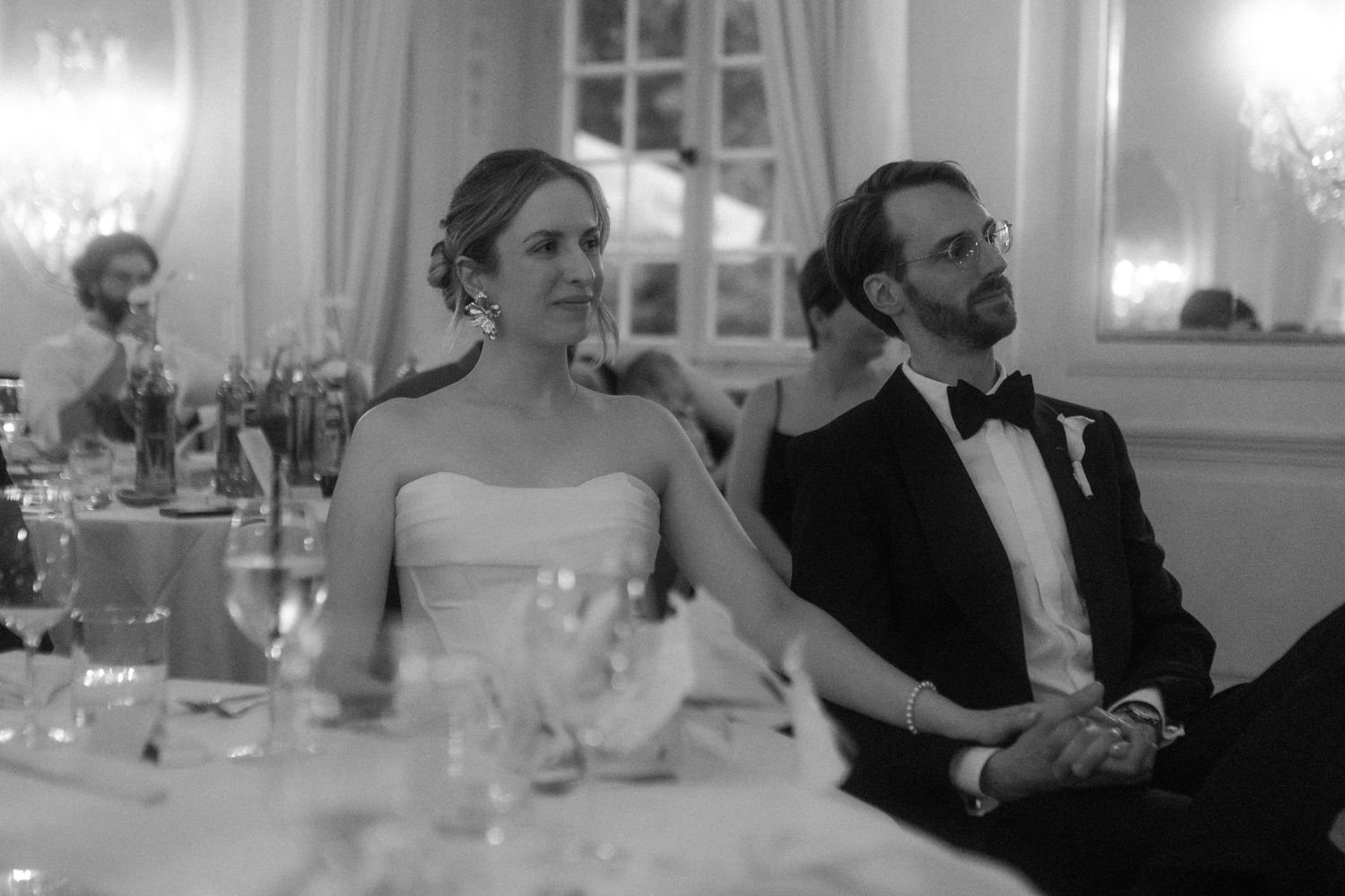 Guests enjoying an elegant wedding reception in a white ballroom with crystal chandeliers.