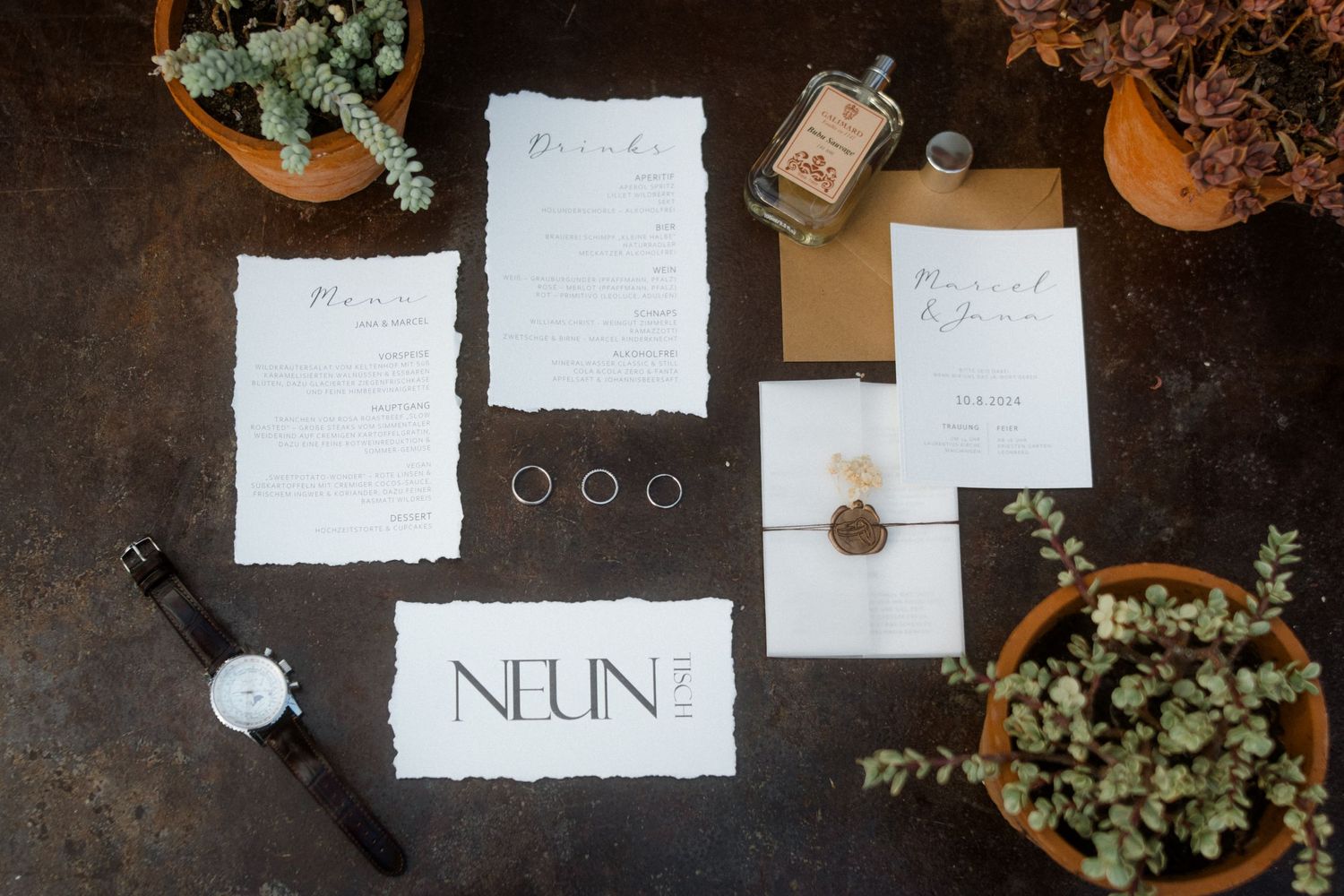 Wedding details including invitation suite, rings, watch, and succulents arranged on rustic surface.