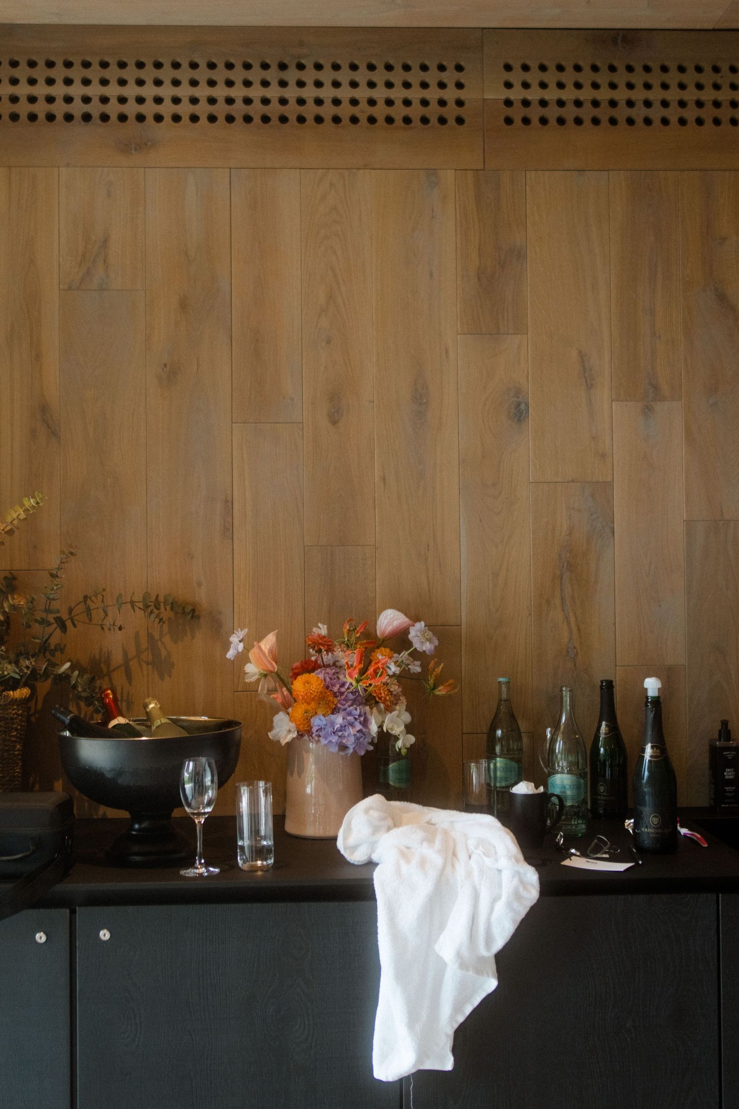 Wedding reception setup with wooden wall, floral arrangement, and wine bottles on dark cabinet.