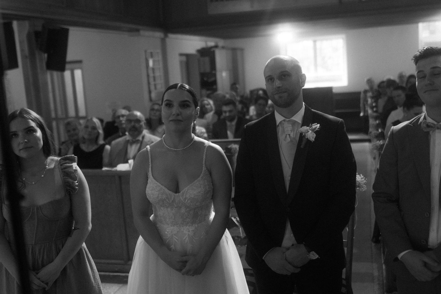 A black and white wedding ceremony photo showing attendees standing in a traditional church setting.