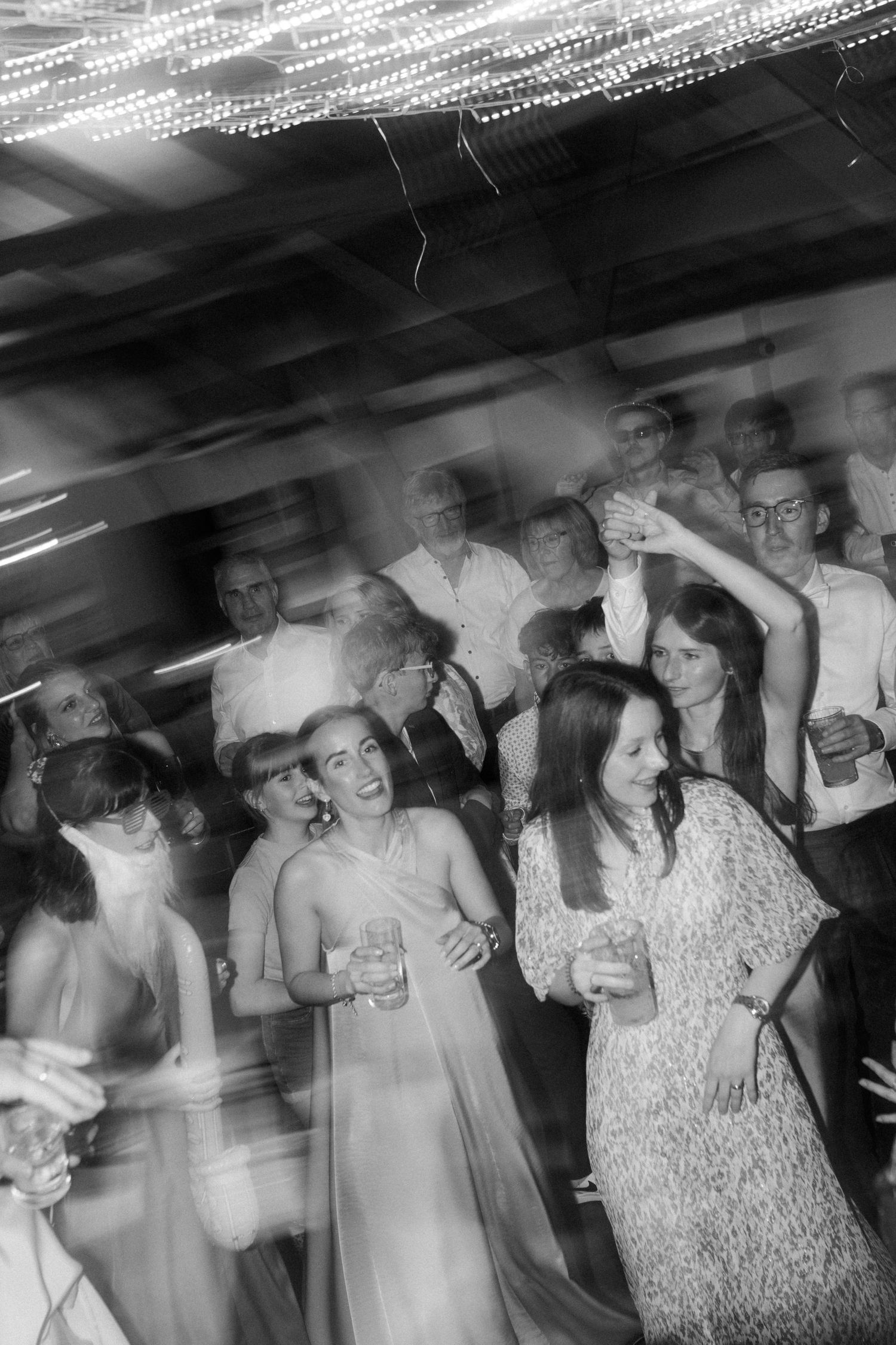 Party guests dancing under string lights in a blurred motion black and white photograph.