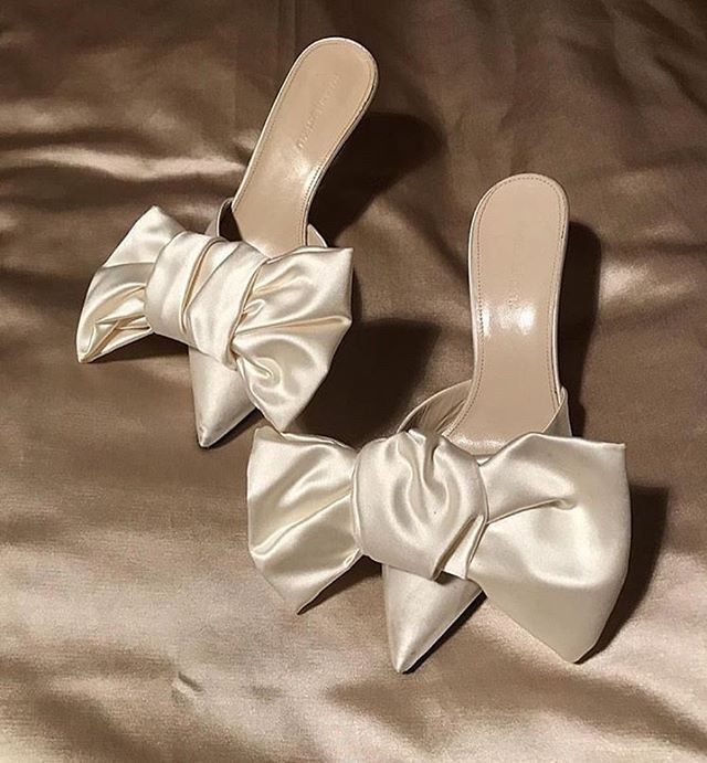 White satin bridal mules with large bow details photographed on silk fabric.