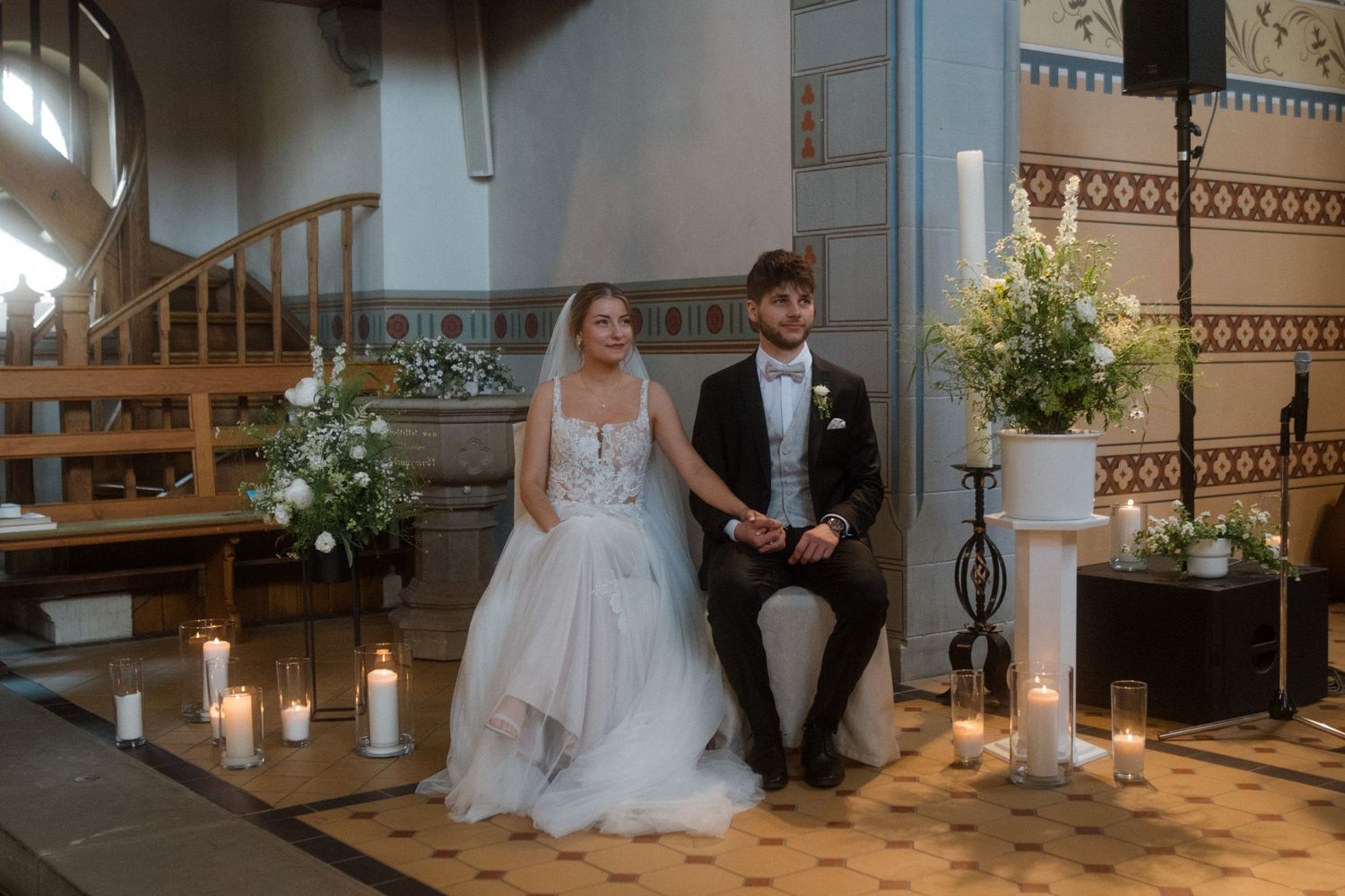 A couple sits surrounded by candles and floral arrangements during their church wedding ceremony.