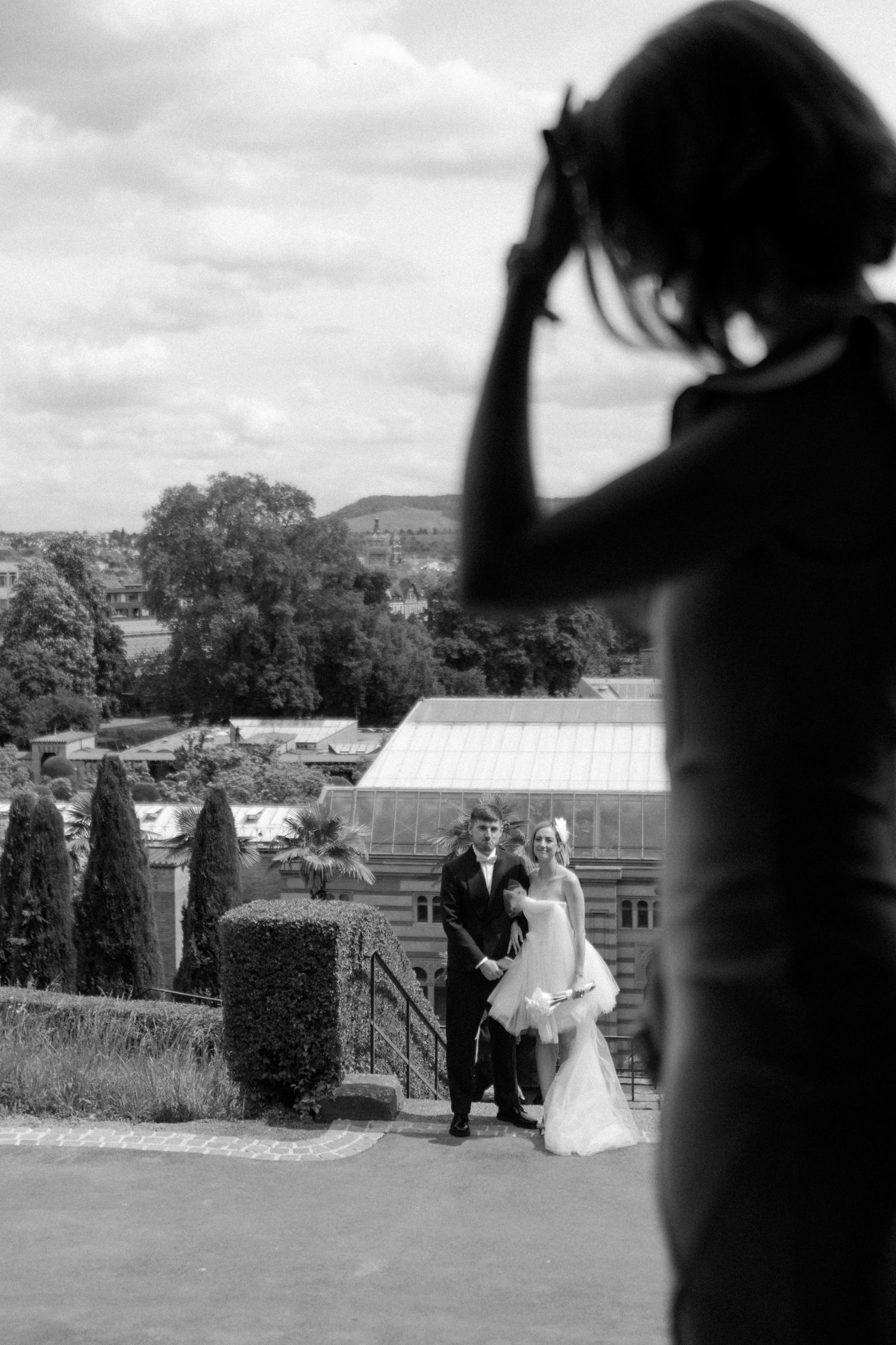 Wedding photography scene at an elegant garden venue with greenhouse in background.