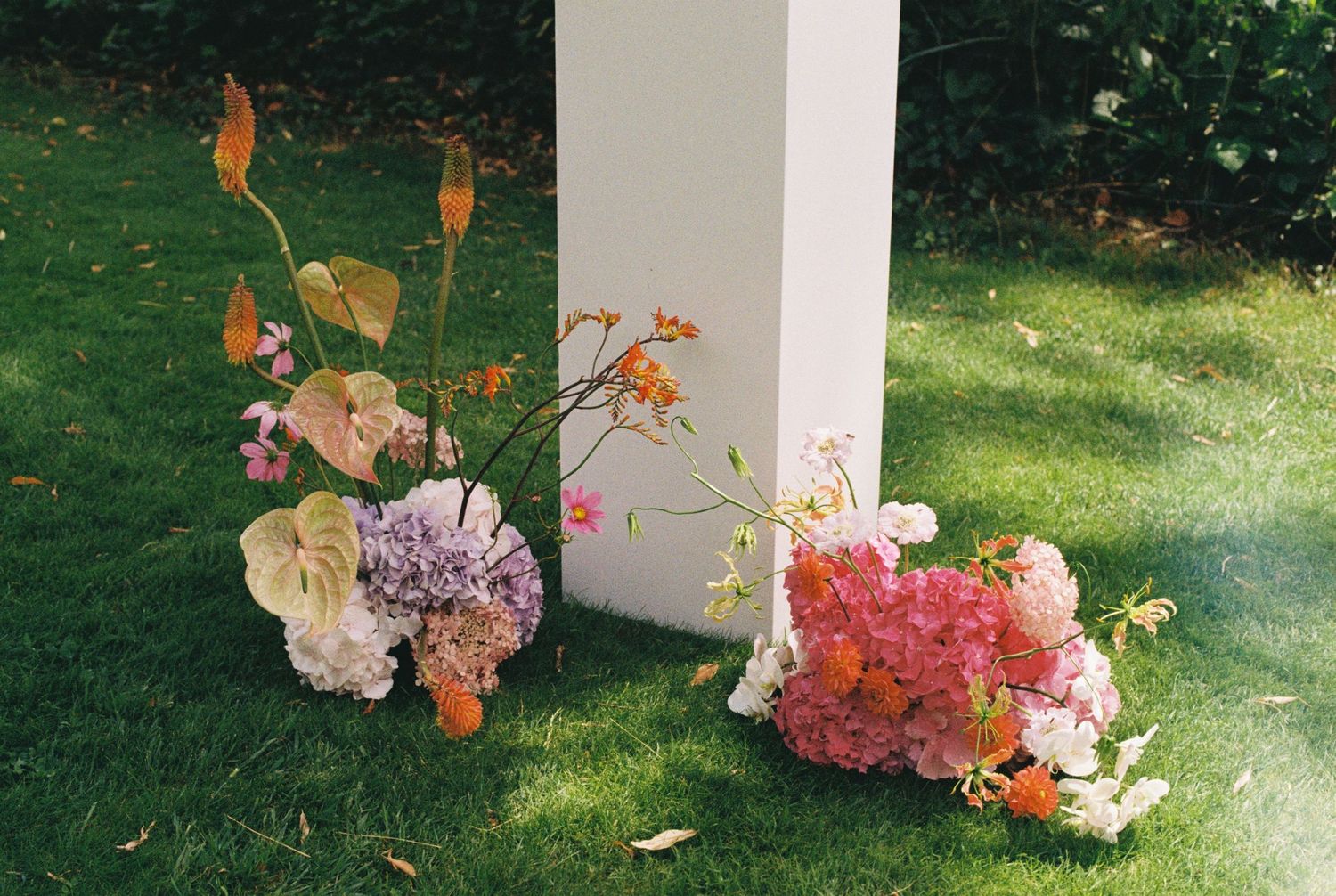 Floral arrangements with pink and white flowers sit at the base of a white column on green grass.
