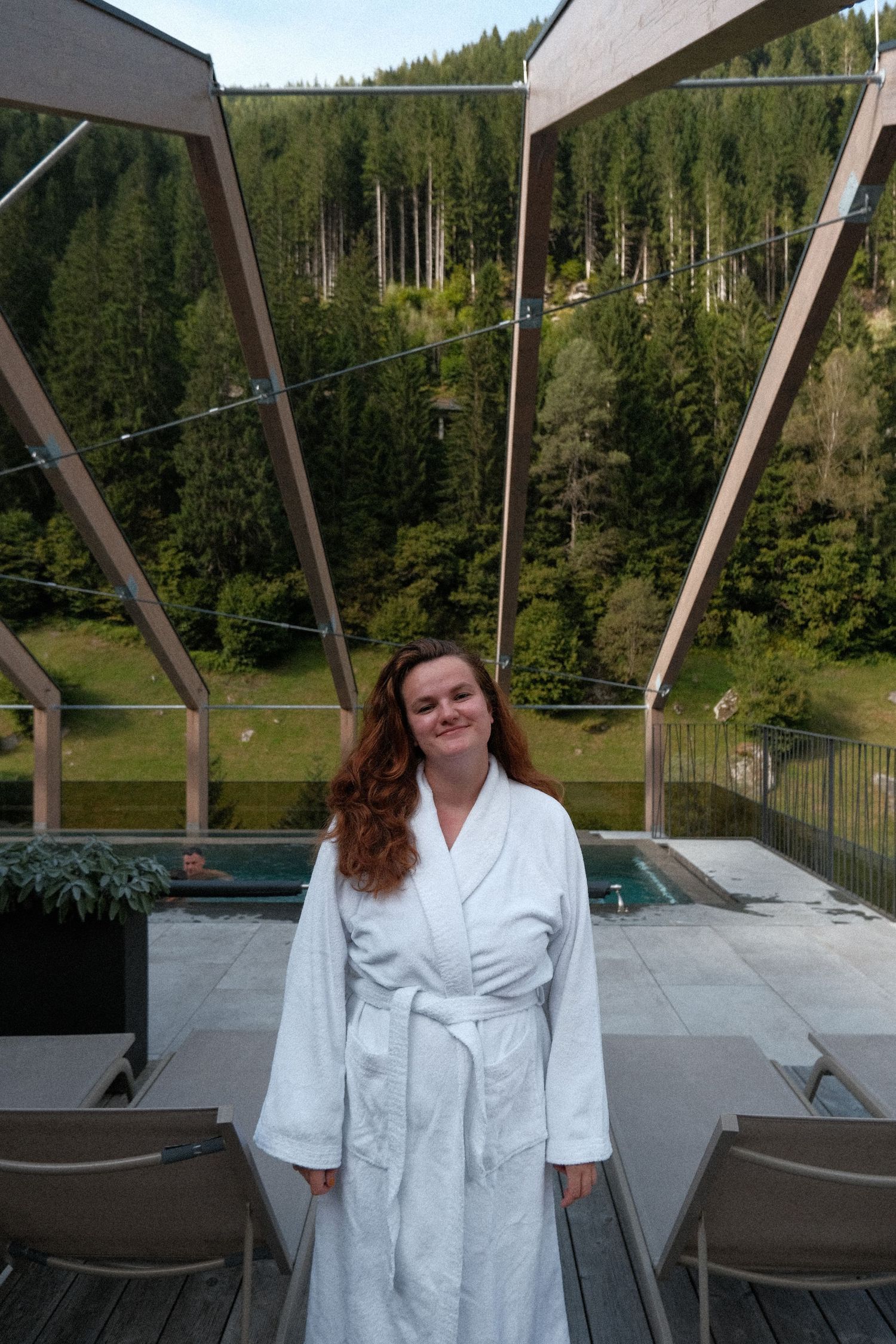 Person in white bathrobe enjoying a mountain view from a spa terrace with lounge chairs.