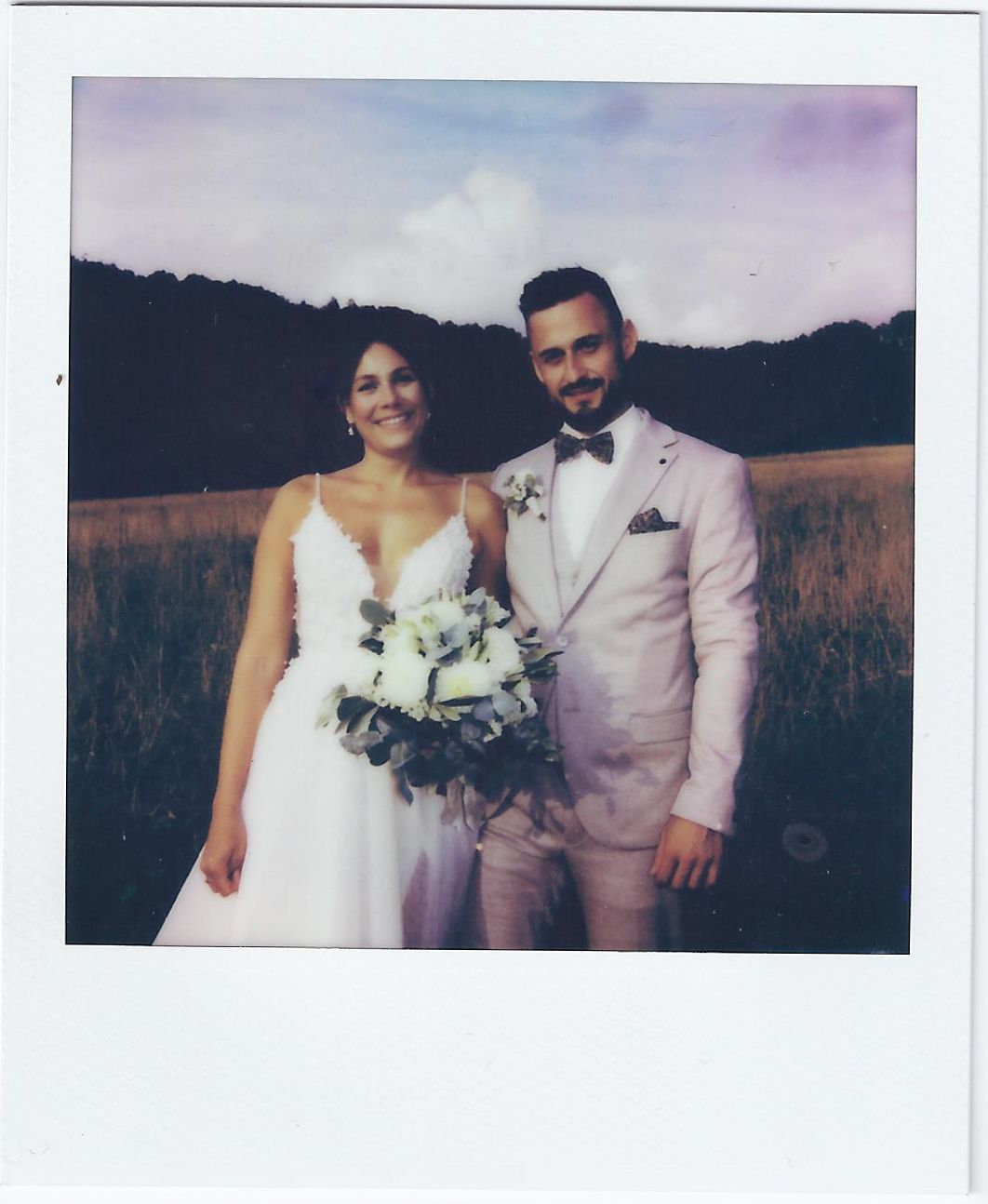 Wedding couple poses together in a field at sunset holding a bouquet of white flowers.