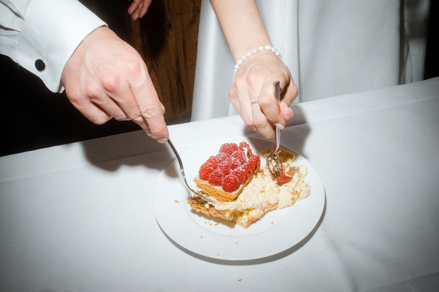 A bride and groom cut a small strawberry-topped wedding cake together on a white tablecloth.