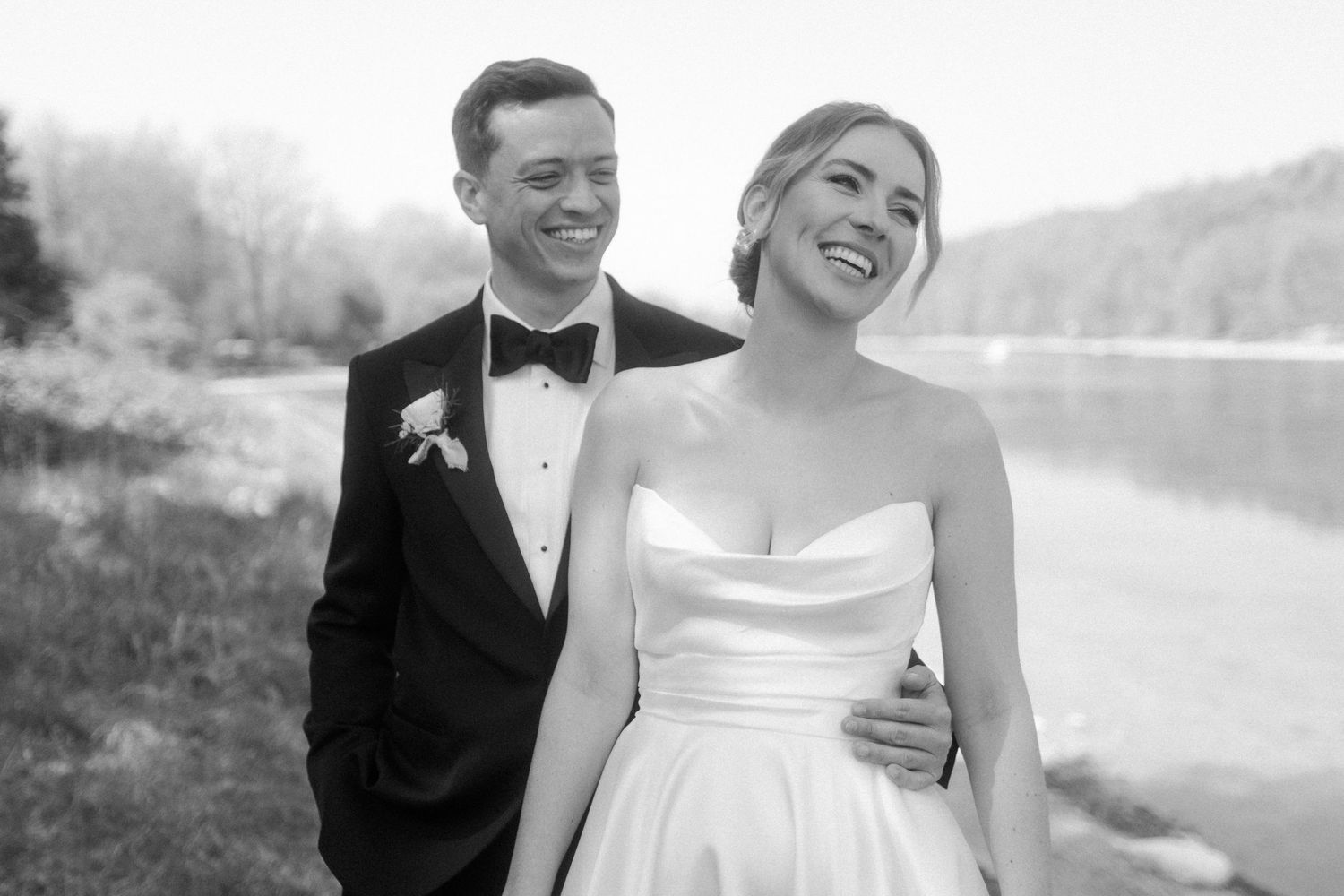 Black and white photo of smiling bride in strapless gown and groom in tuxedo posing by a lakeside.