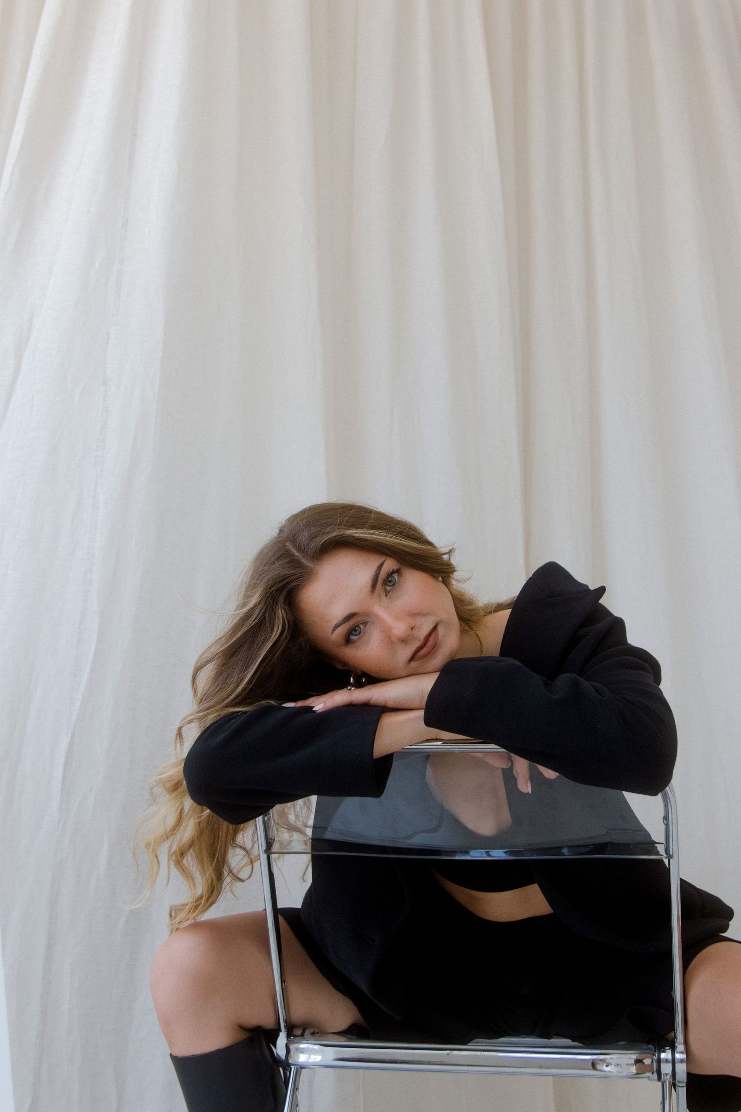 Young woman in black outfit resting chin on hands atop clear chair against white draped backdrop.