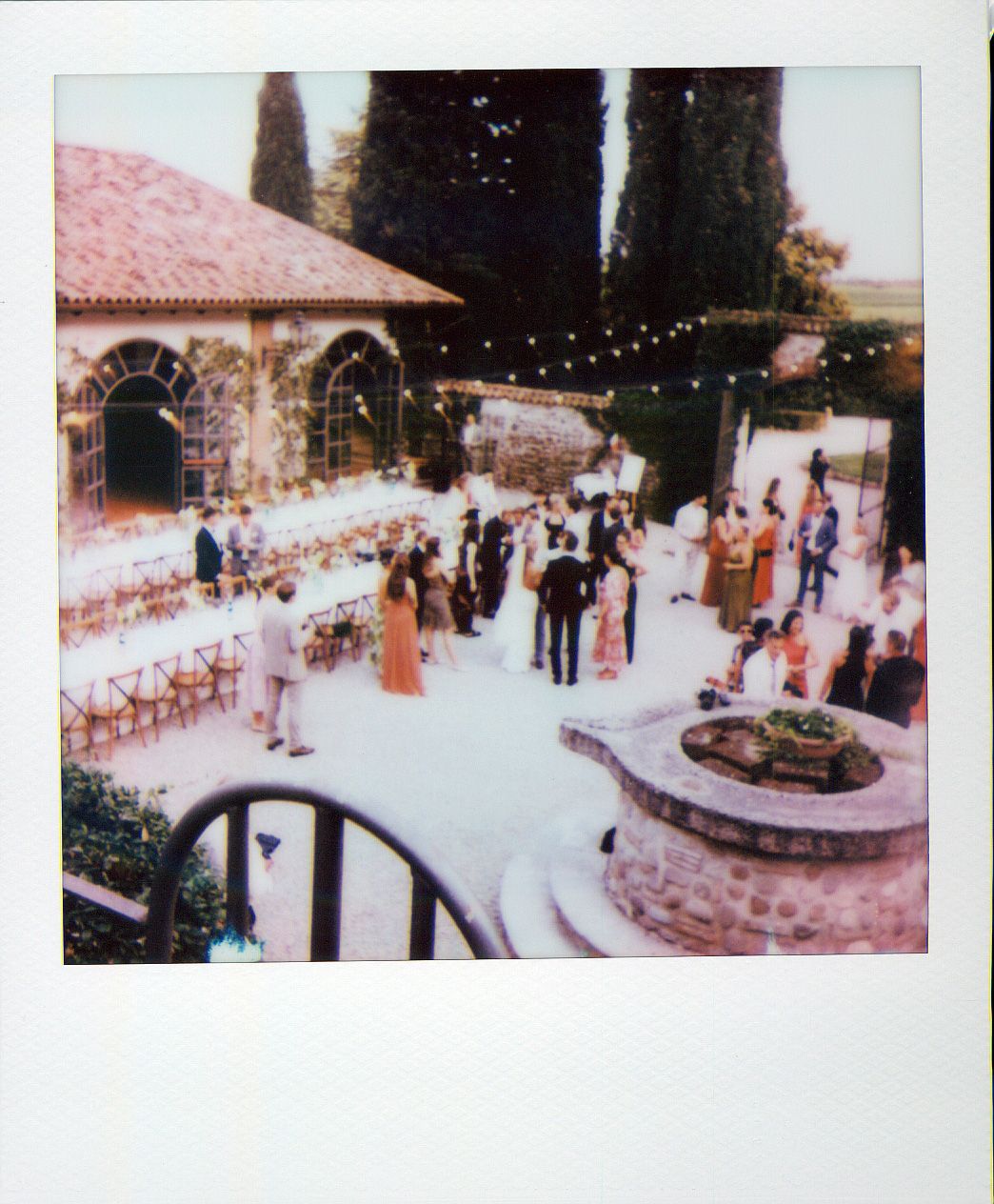 Guests gather at an outdoor wedding reception venue with string lights and a stone fountain centerpiece in a Mediterranean setting.