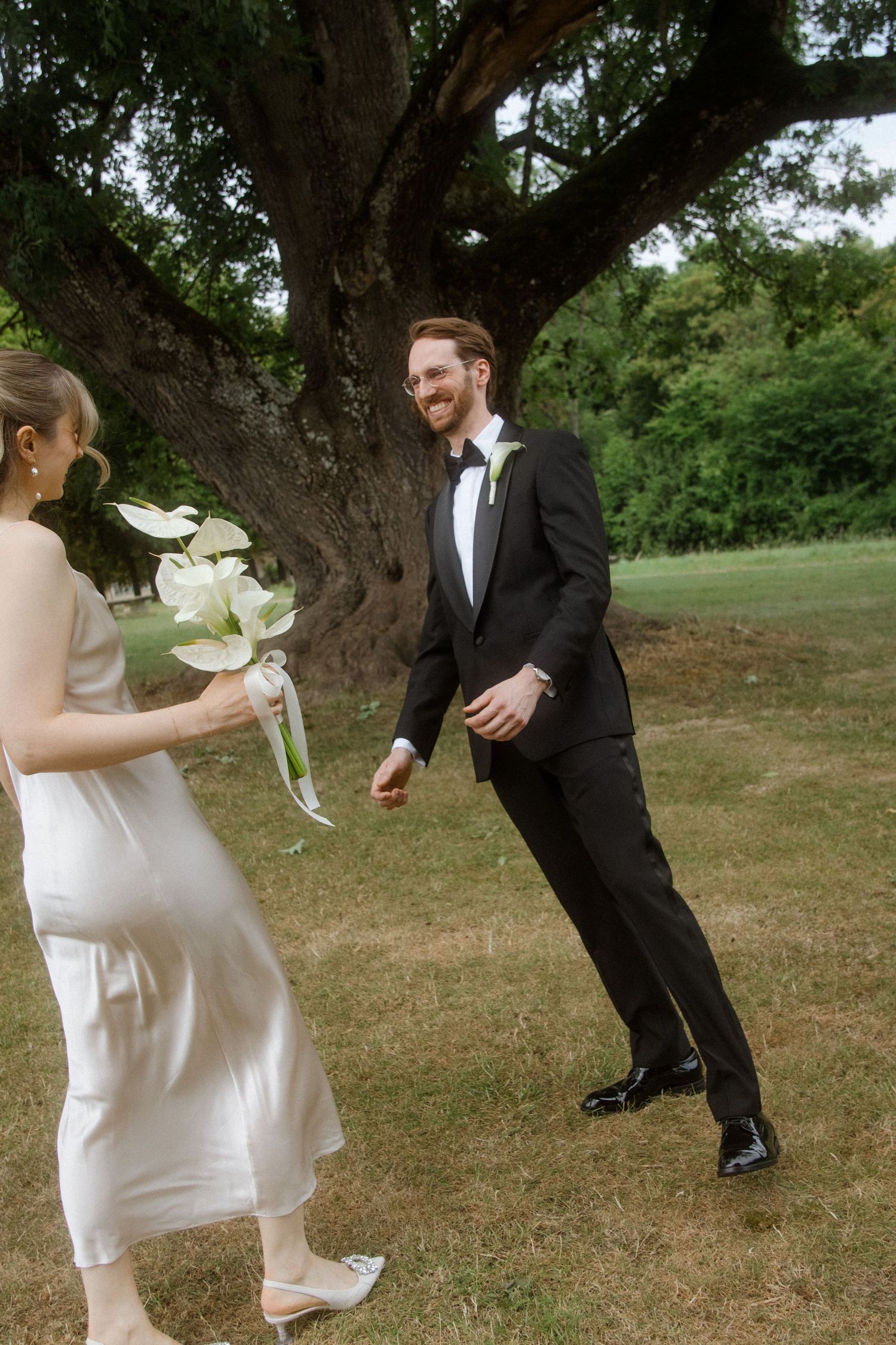 A couple shares a playful moment outdoors under a large tree during a wedding photoshoot.