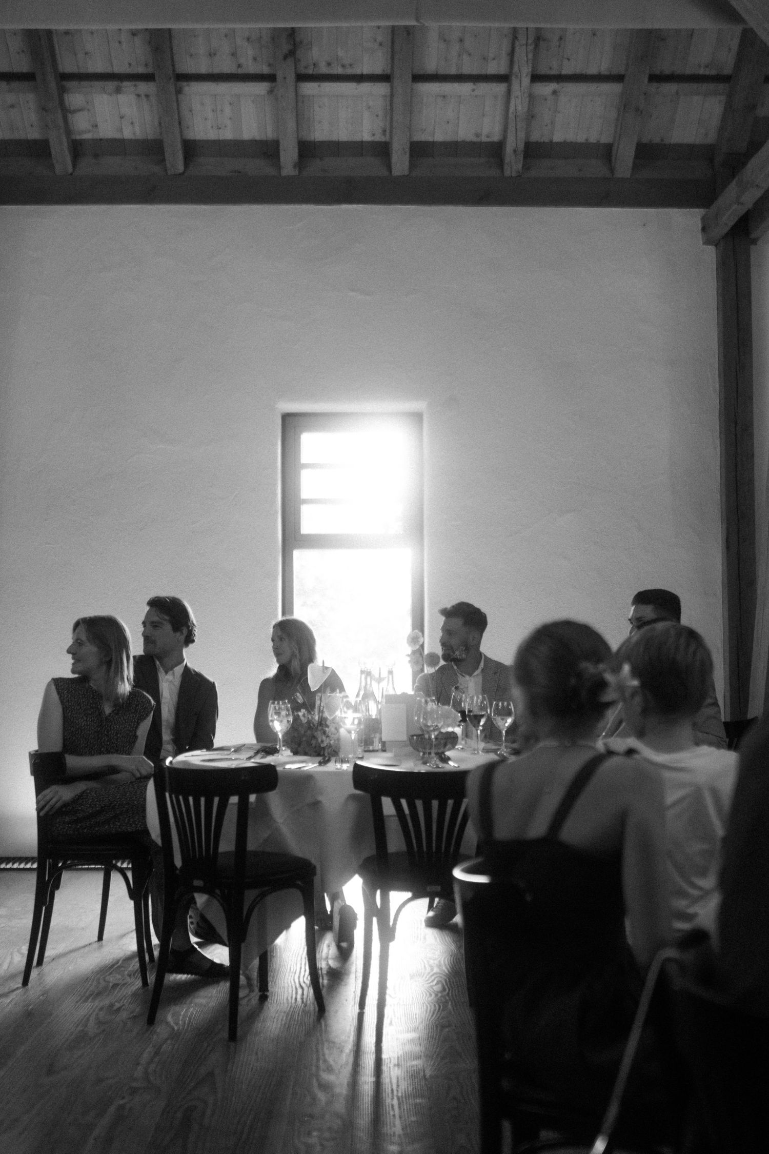 Black and white photograph of wedding guests seated at tables near bright window.