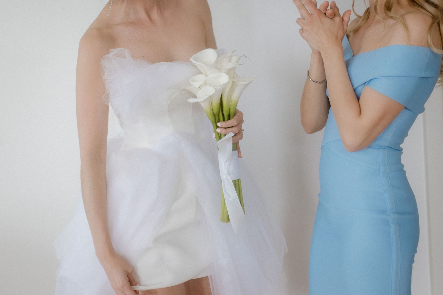 Two women in white and blue dresses holding a bouquet of white calla lilies.