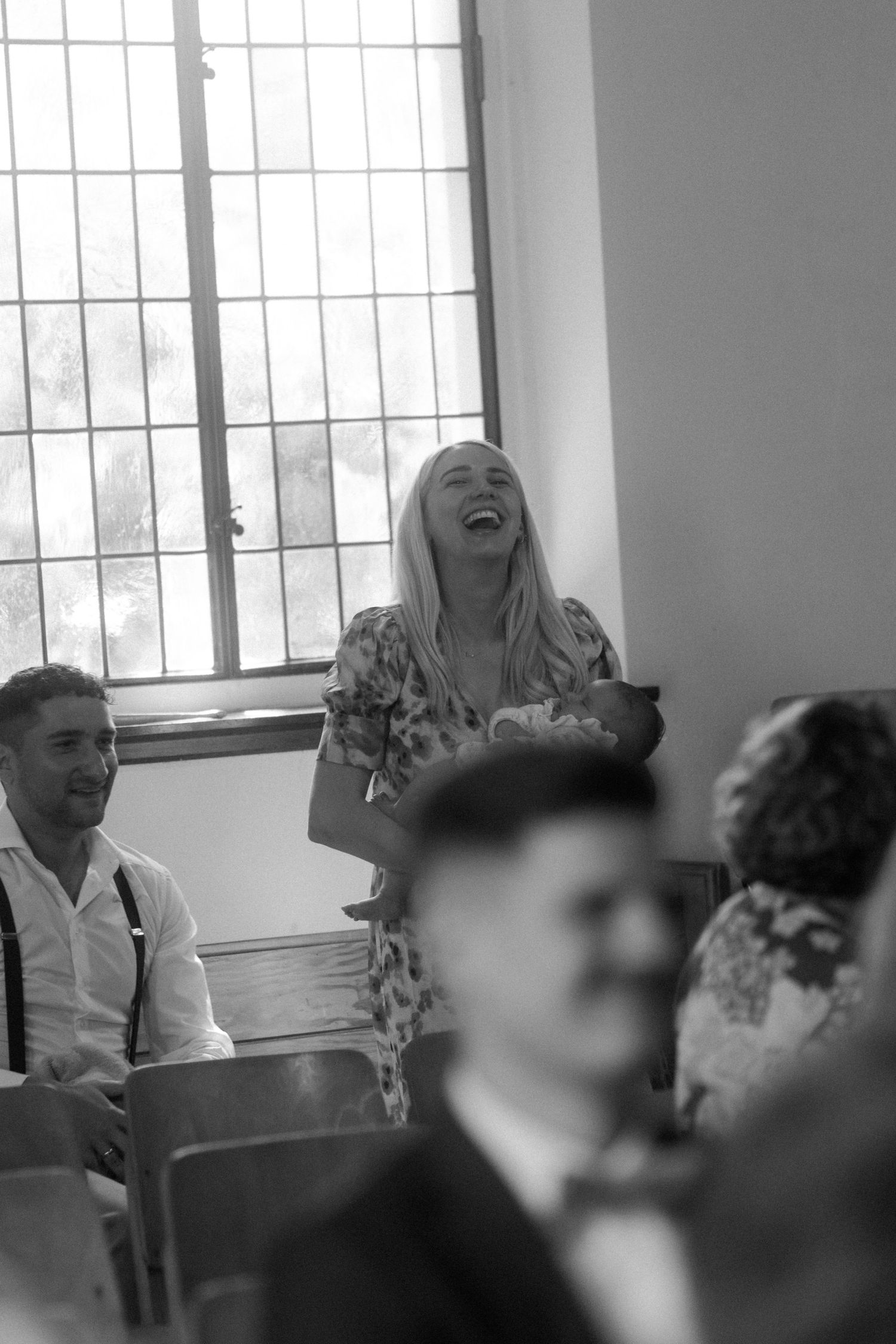 A black and white photo of wedding guests laughing during the ceremony near a bright window.