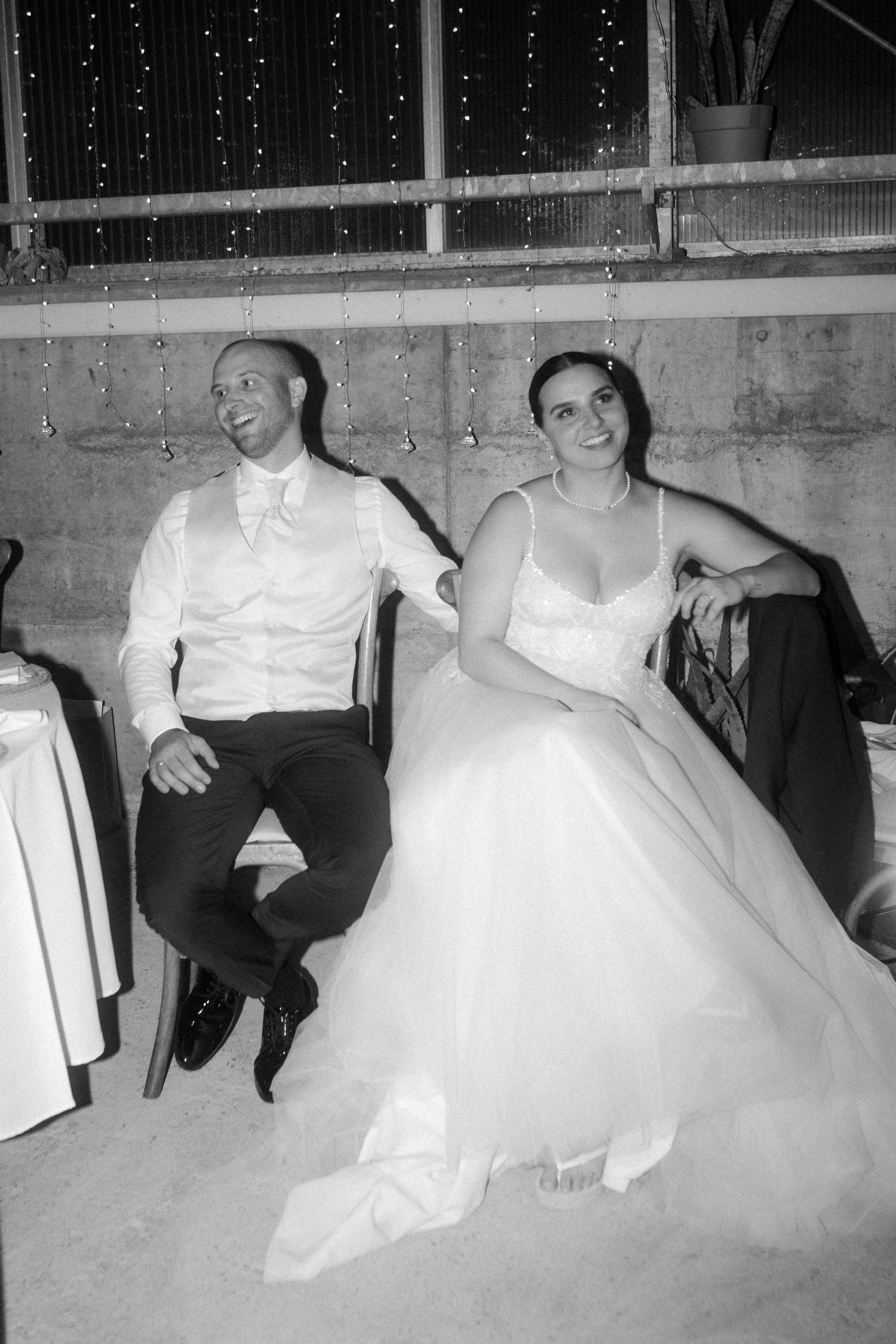 Wedding couple seated and laughing during reception under decorative string lights.