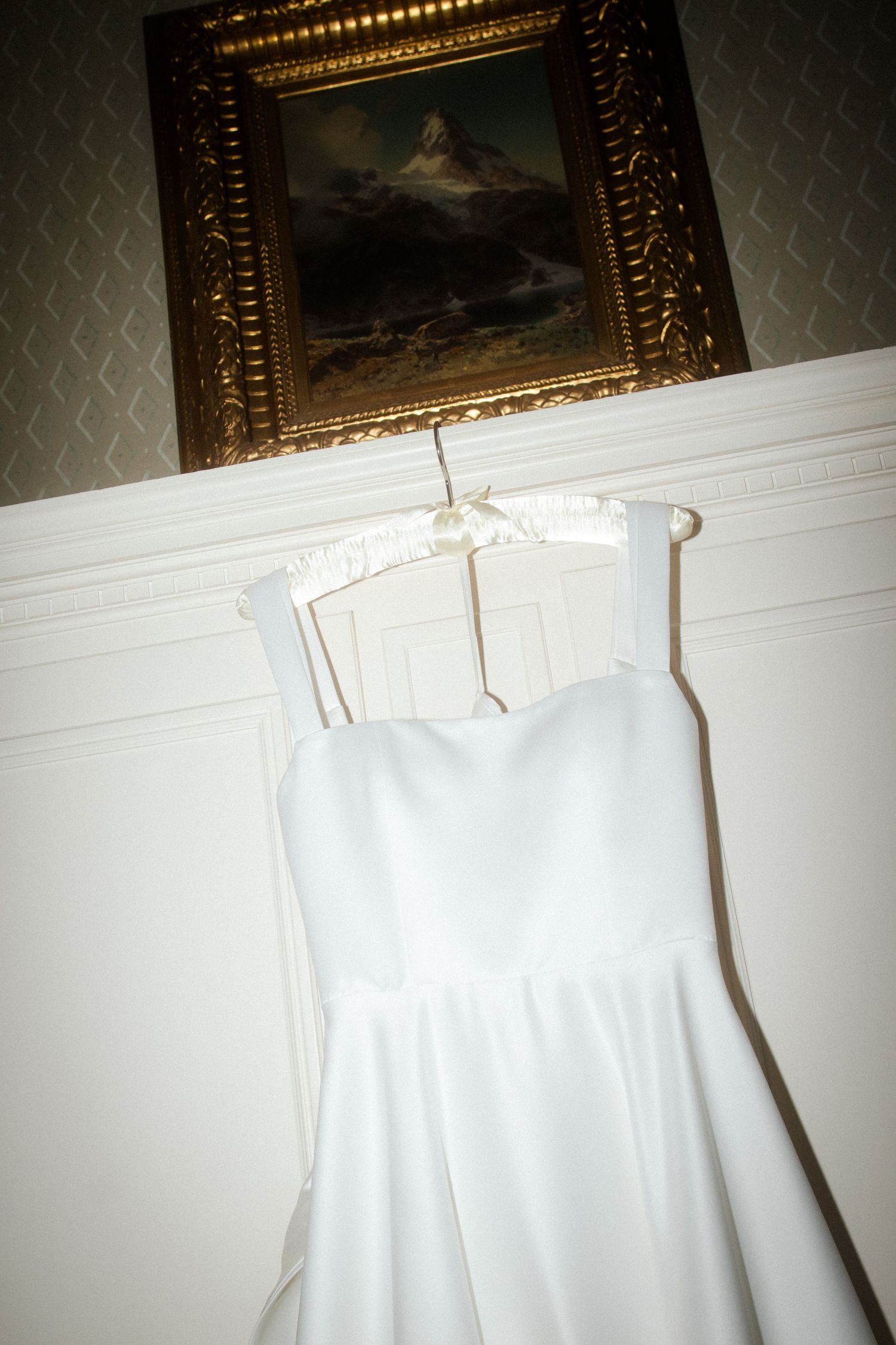 White wedding dress hanging against wall with ornate gold-framed artwork above.
