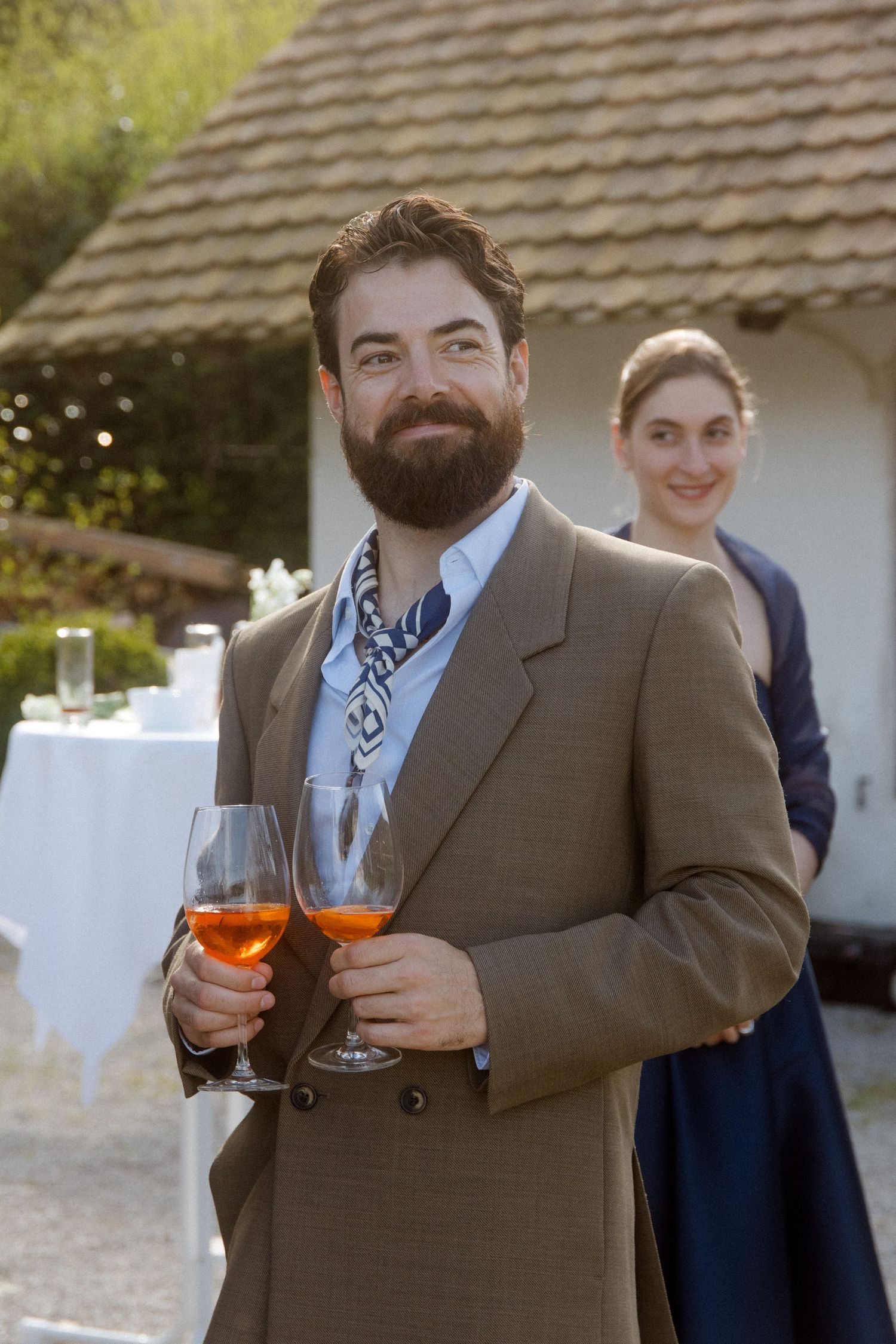 Bearded man in tan suit holding rose wine glasses smiling at an outdoor wedding reception.