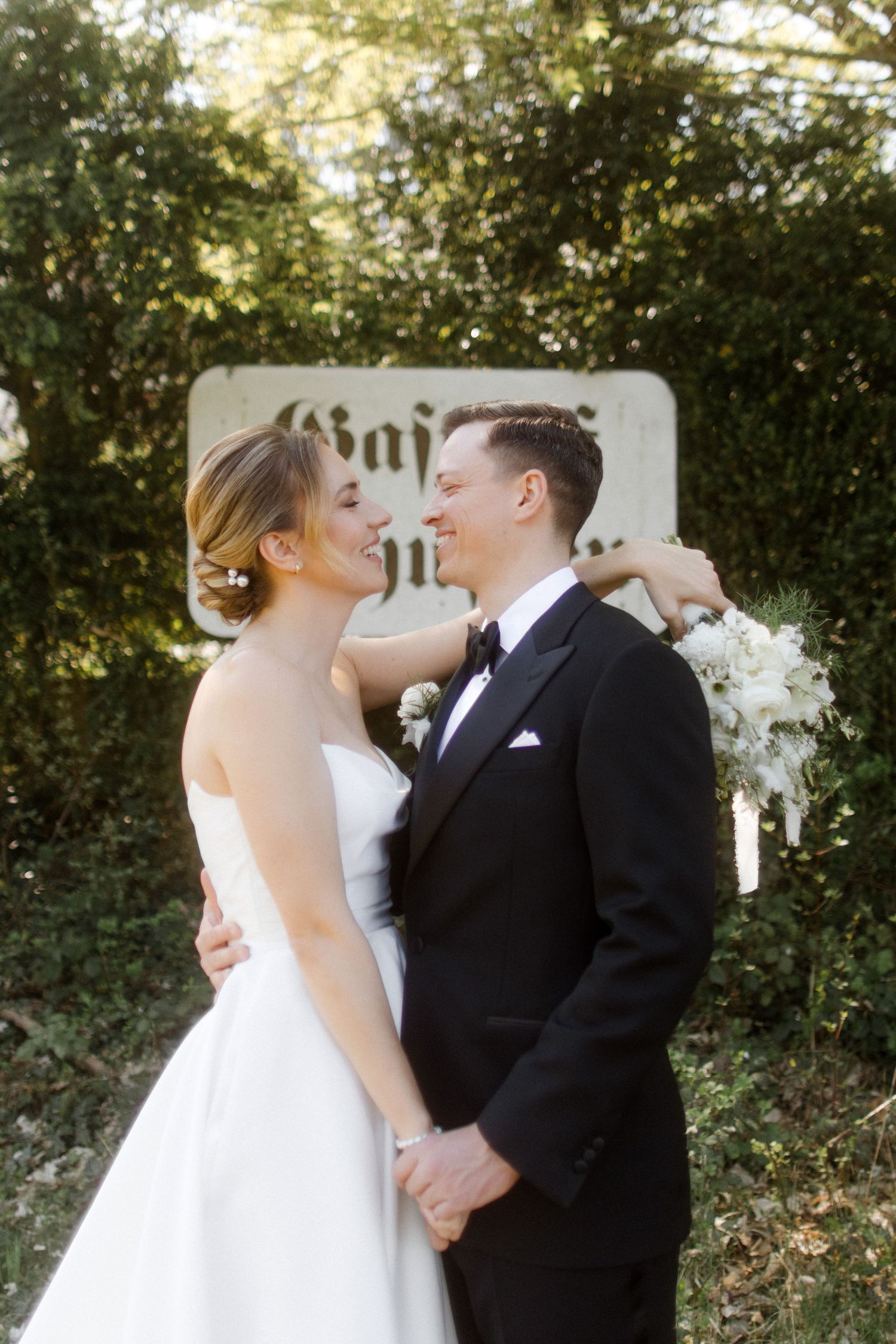 Bride and groom smiling at each other in wedding attire, holding hands and white bouquet before a stone sign outdoors.
