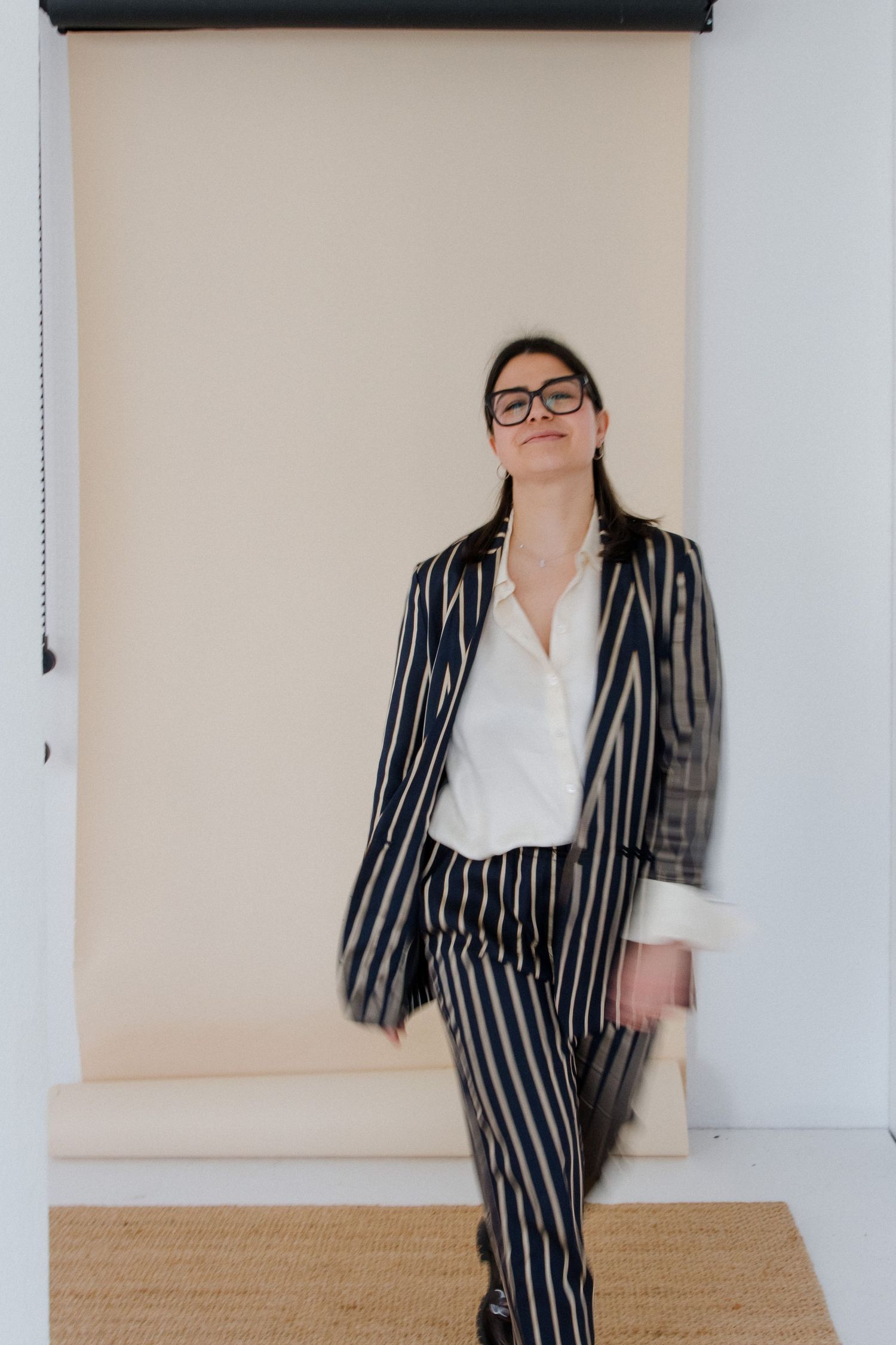 Woman in striped navy suit and white shirt poses confidently against beige backdrop wearing black glasses.