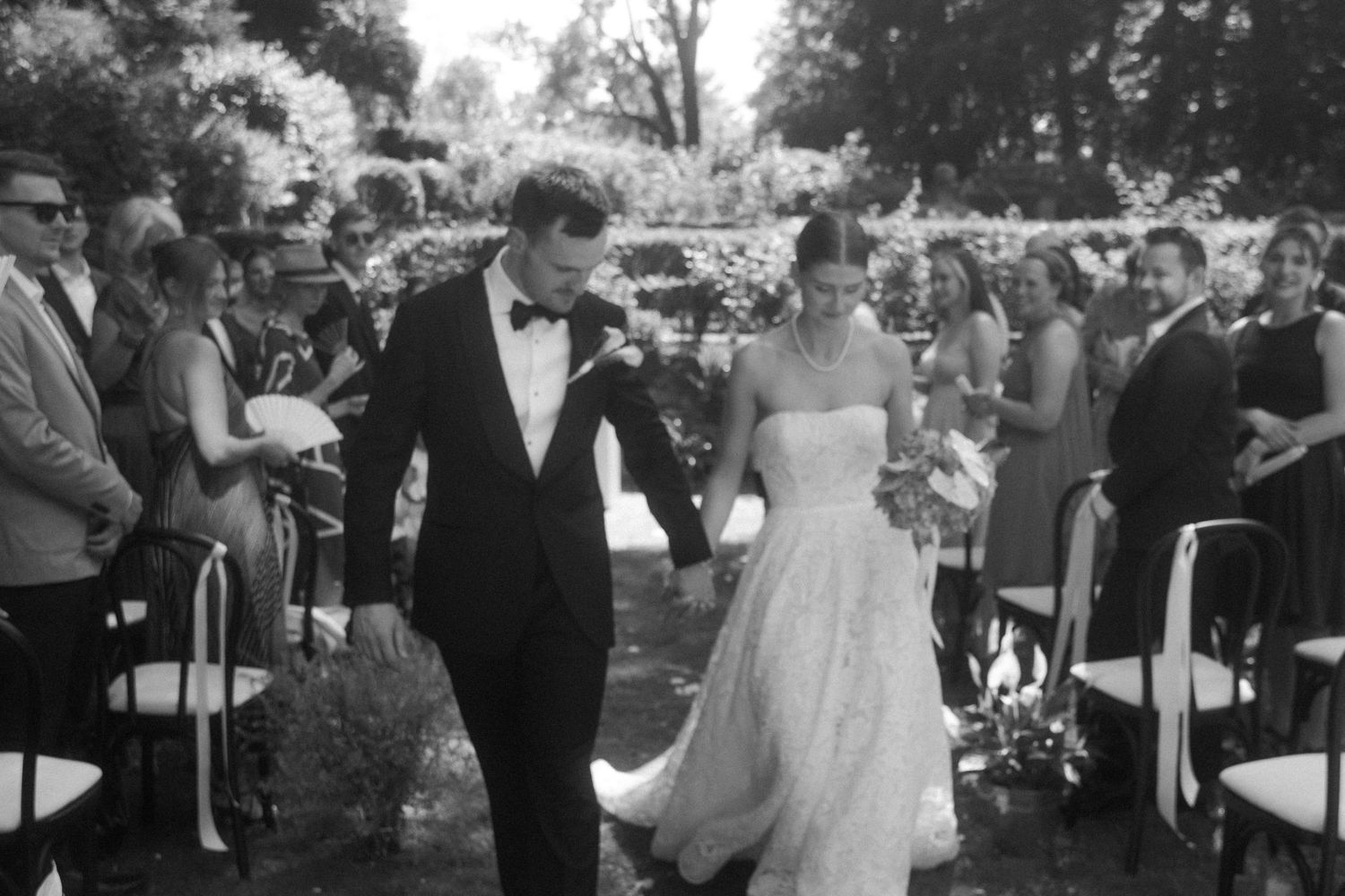 Black and white photo of a bride and groom walking down the aisle at an outdoor wedding ceremony.
