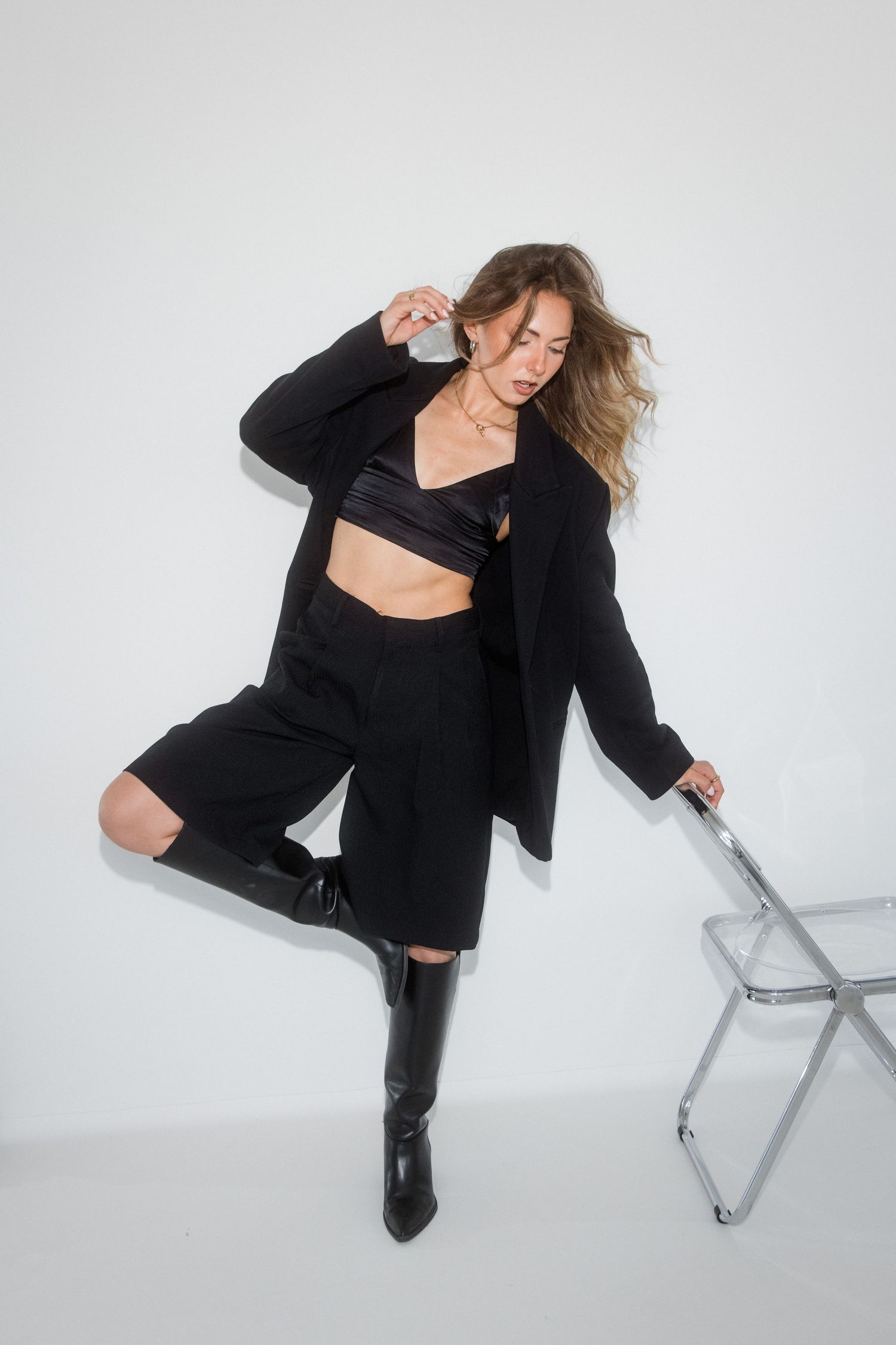 Woman in black blazer, crop top, shorts, and knee-high boots posing dynamically beside a clear chair.