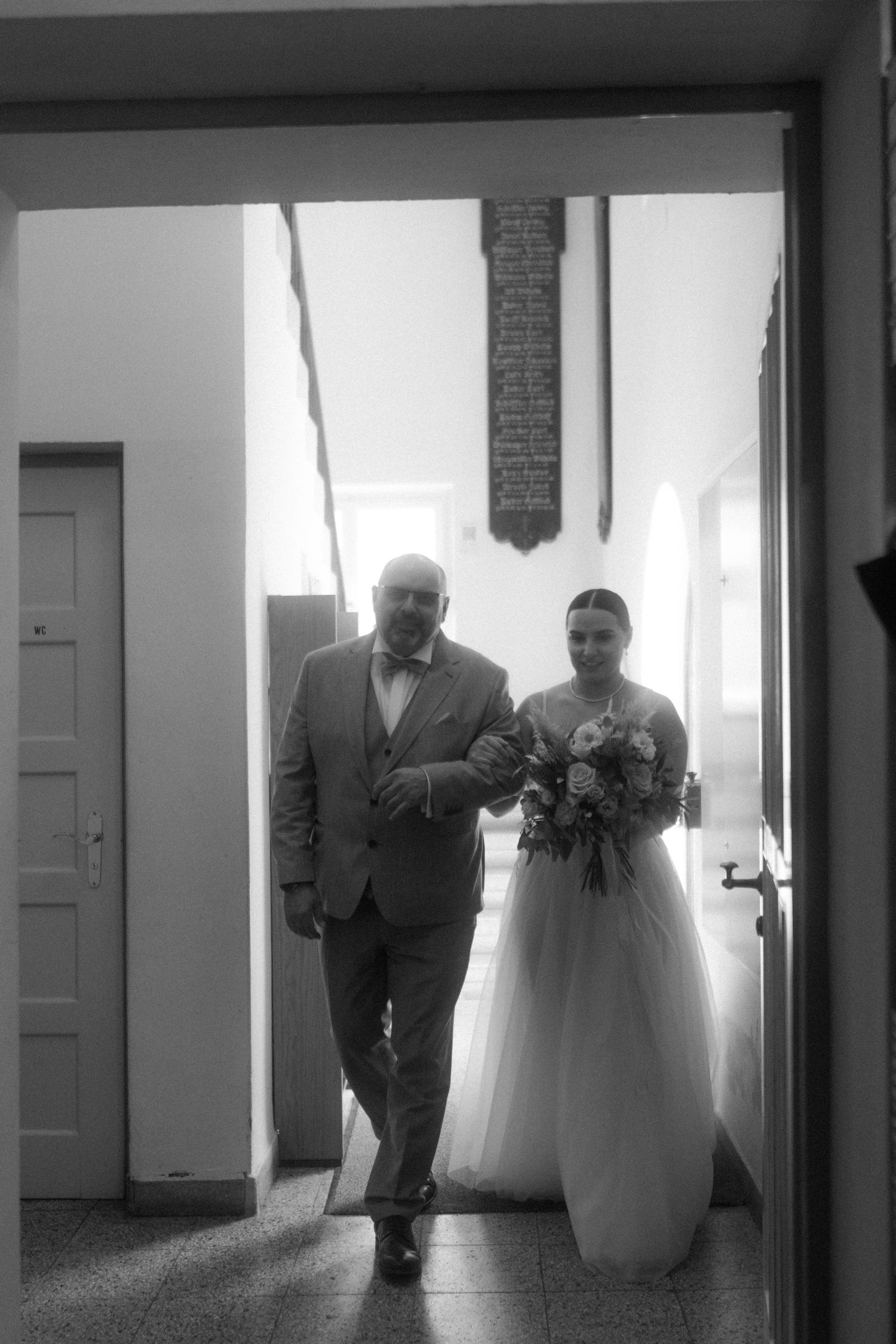 A silhouetted couple walks down a bright hallway in formal wedding attire.