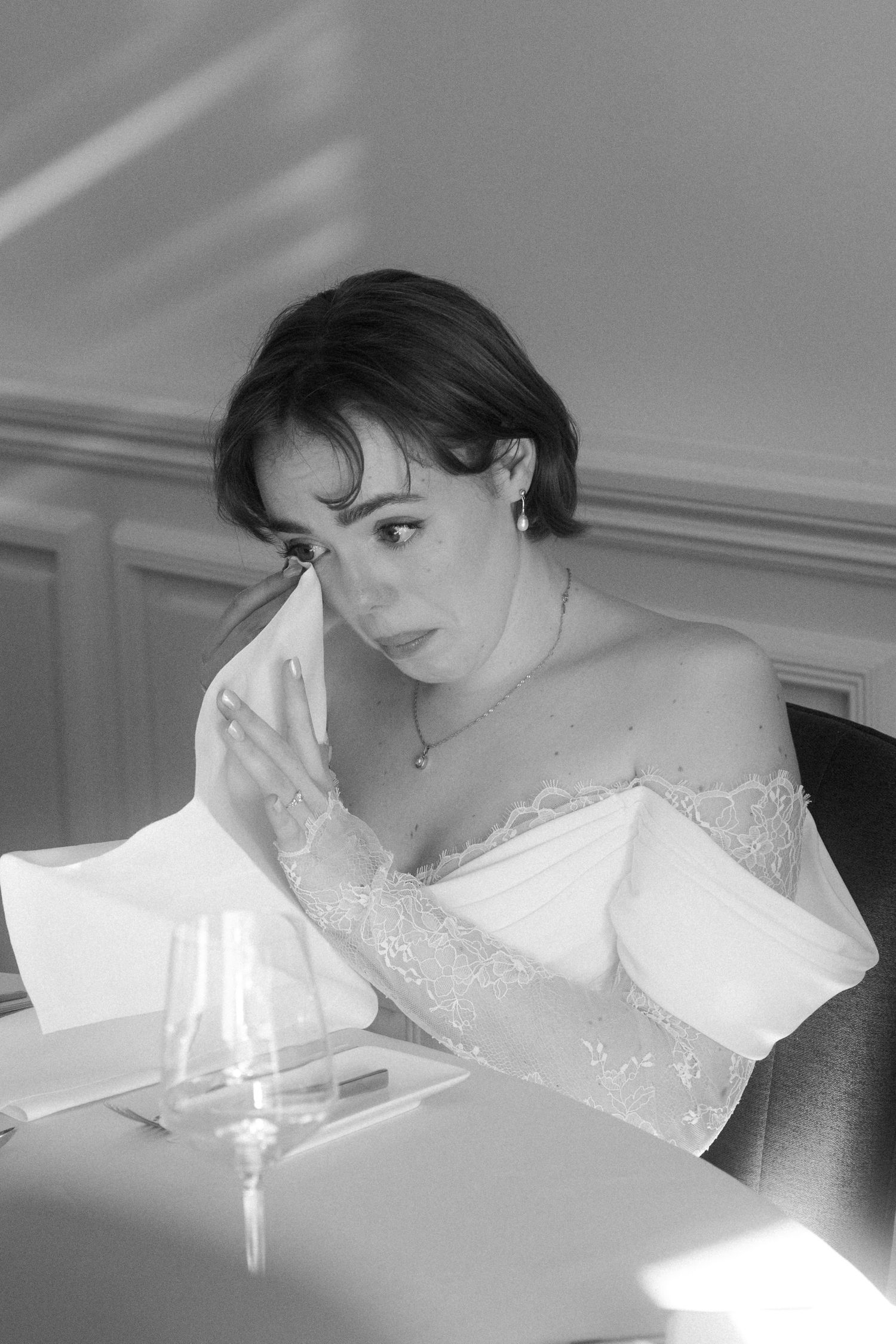 Black and white photo of an elegant woman in off-shoulder lace dress sitting at a dining table with a wine glass.