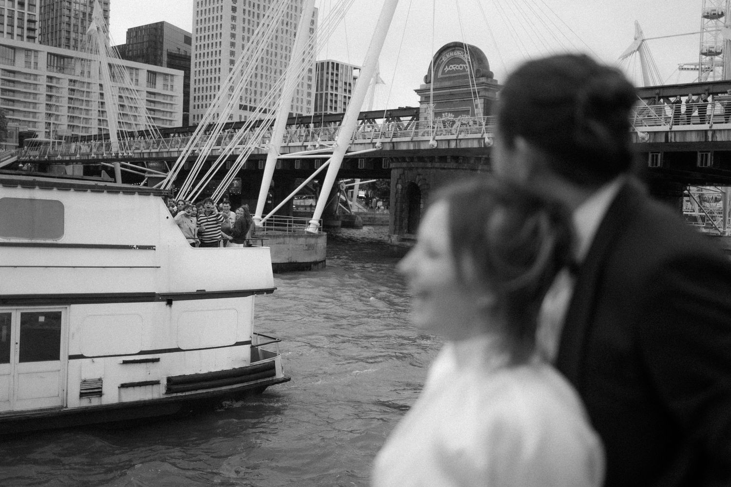 Black and white photo of a couple in wedding attire by a river with a boat and bridge in the background.