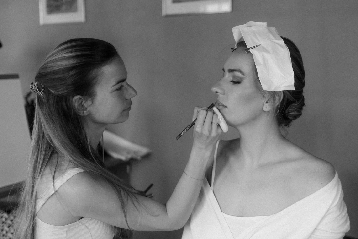 Makeup artist applying lipstick to a bride in a white dress and hair wrap, black and white photo.