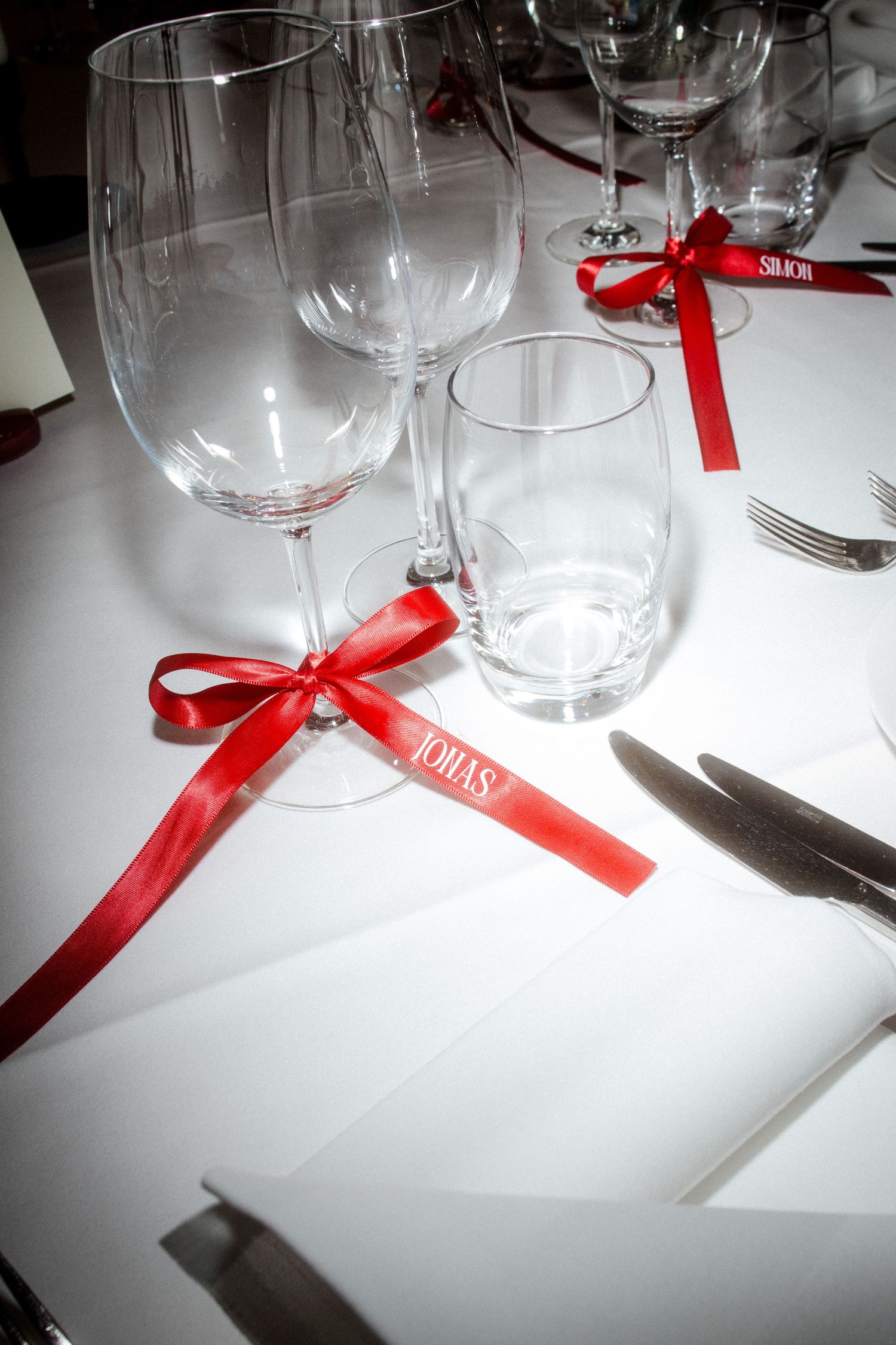 Close-up of wedding place setting with crystal glasses adorned with red ribbon name tags on white tablecloth.