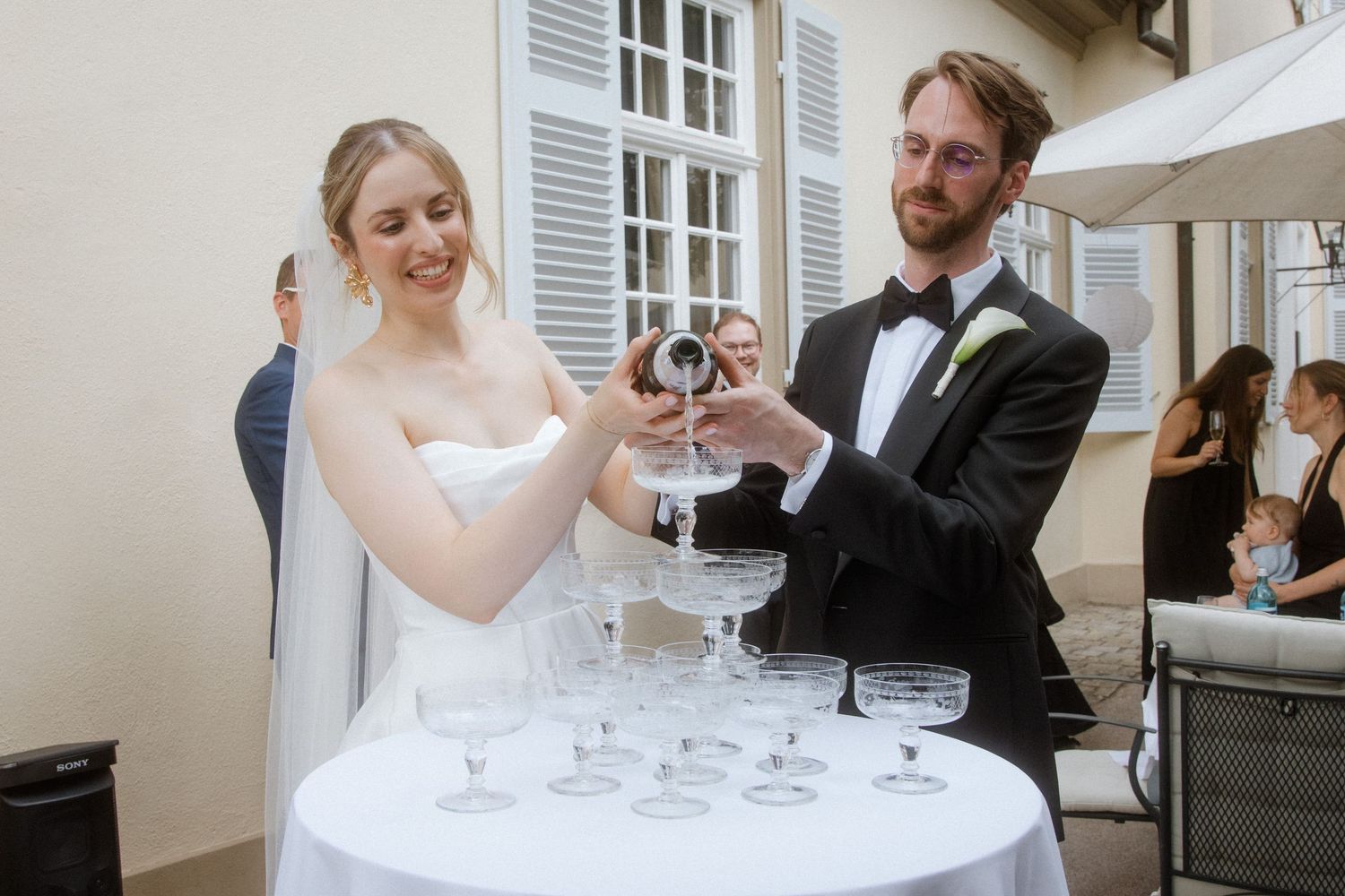 A couple pouring champagne into a tower of glasses at an outdoor wedding reception.