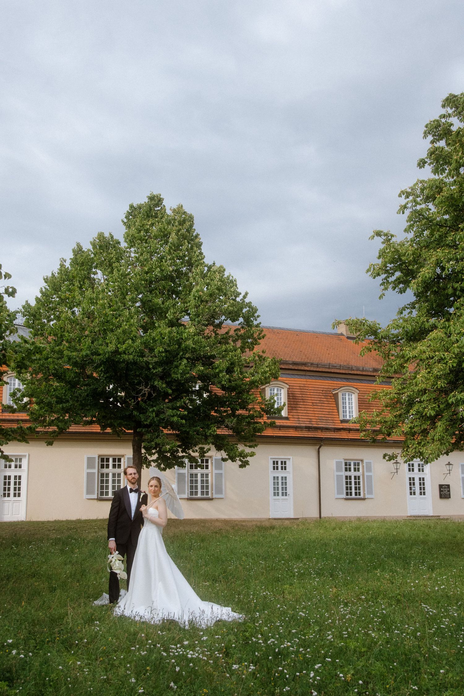 Wedding couple poses against a historic building with orange roof in a grassy meadow.