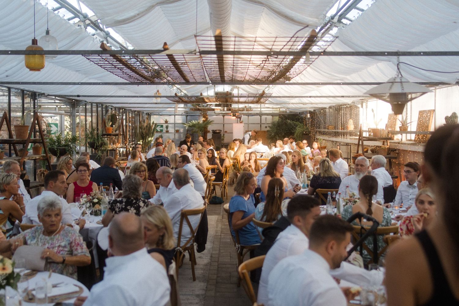Wedding reception guests seated at long tables under a covered outdoor venue with string lights.