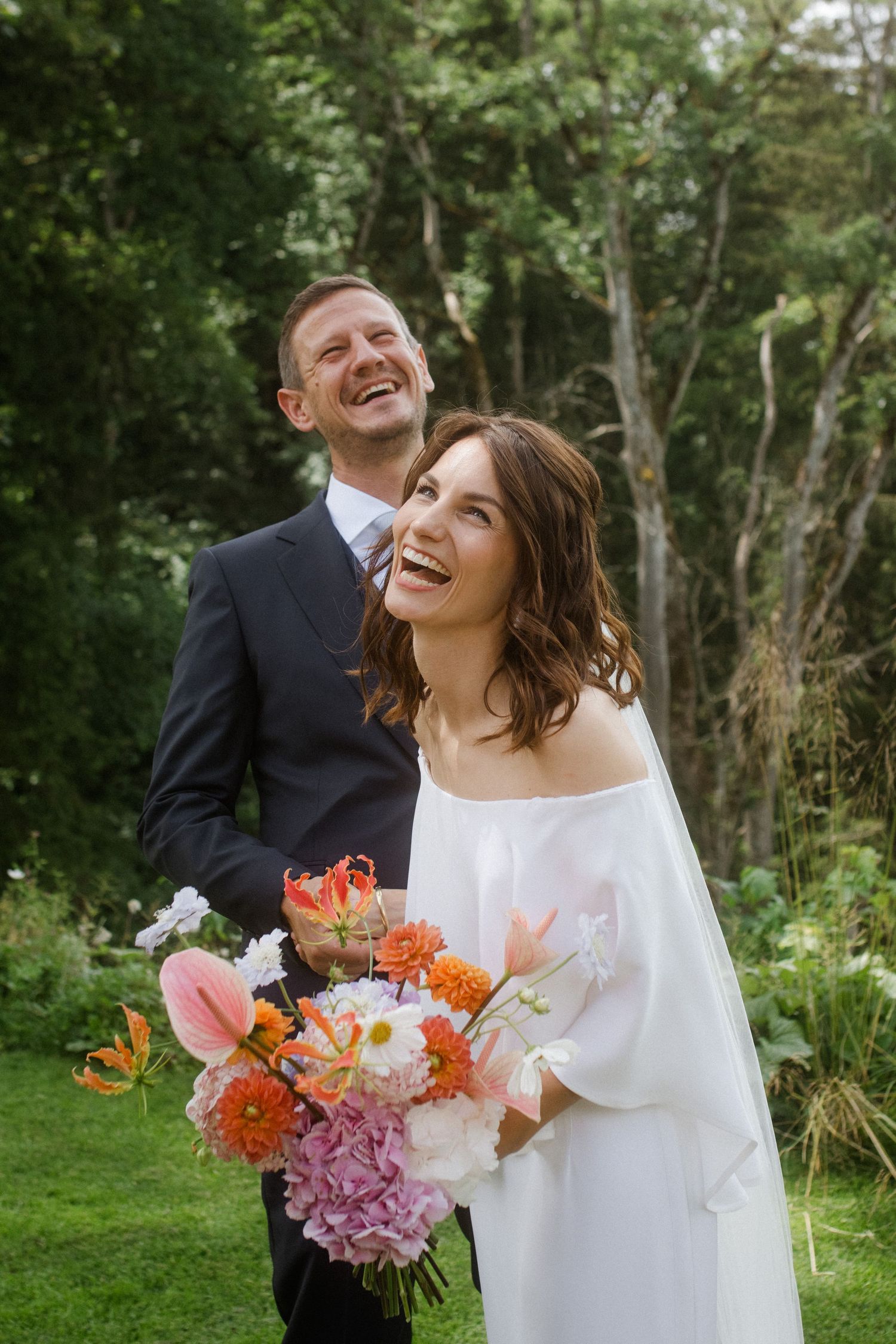 A couple shares a candid laughing moment while holding a bright coral and pink bouquet.