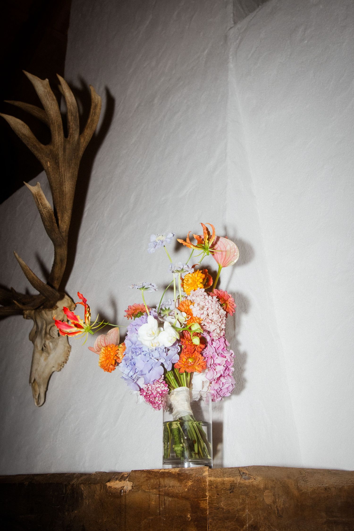 A rustic floral arrangement with orange and purple blooms next to an antler wall decoration.